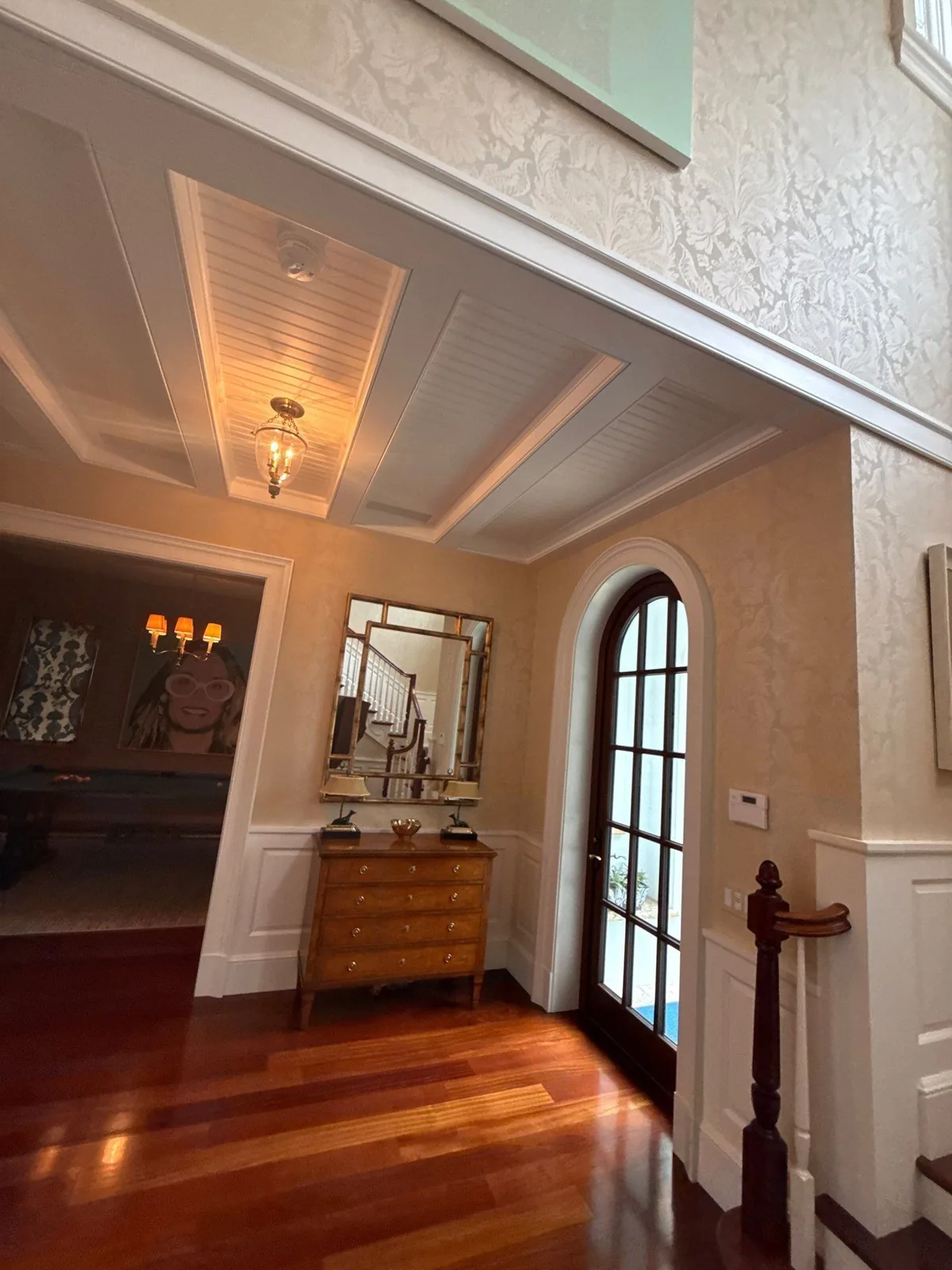 Interior Painting in Palm Beach, Florida. Entry hall with a white painted beadboard and beam ceiling, textured cream walls, white trim, an arched glass door and dark hardwood floor, with a chest of drawers and mirror against the wall.