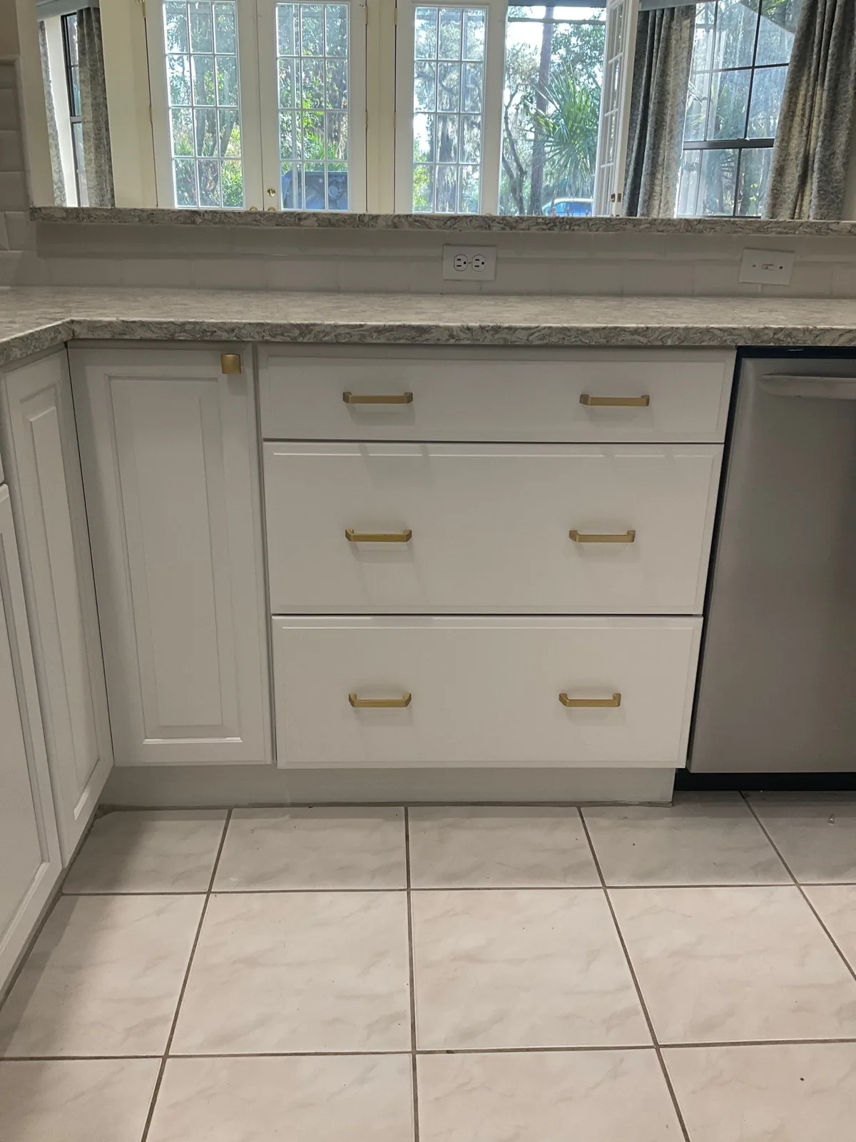 Cabinet Refinishing in Palm City, Florida. Refinished white cabinet drawer bank with long brass pulls below a stone countertop and window.