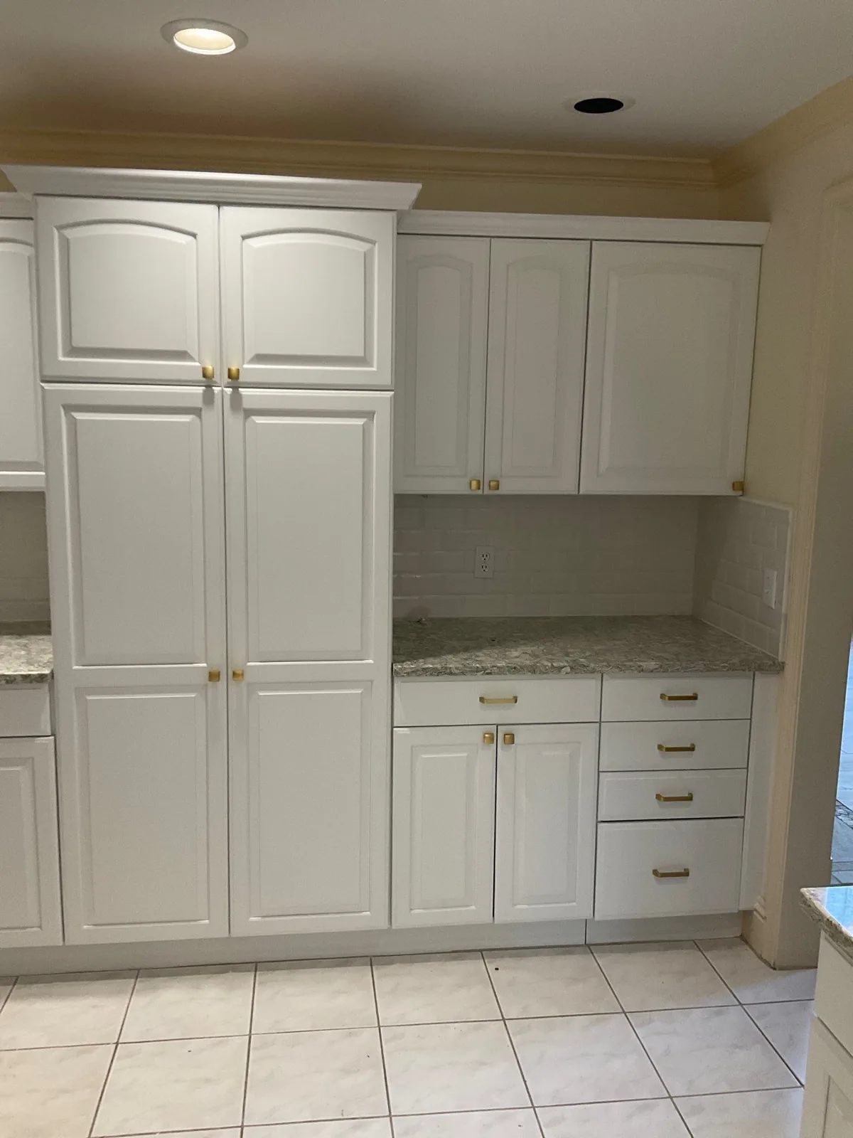 Cabinet Refinishing in Palm City, Florida. Finished white raised-panel kitchen cabinets with tall pantry unit, gold knobs and pulls, speckled granite countertop and tile floor.