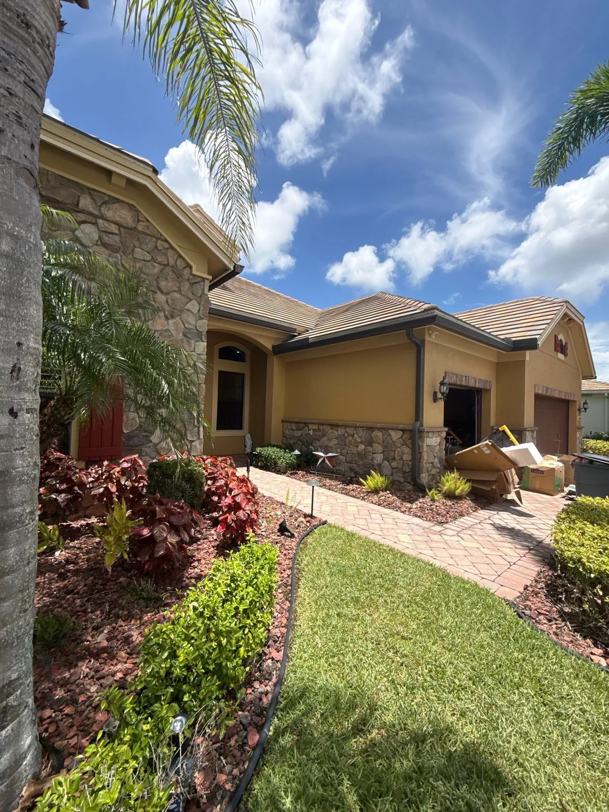 Exterior Painting in Palm City, Florida. Single-storey stucco house exterior in tan with stone veneer accents, tile roof, red front door, paver walkway and planted beds.
