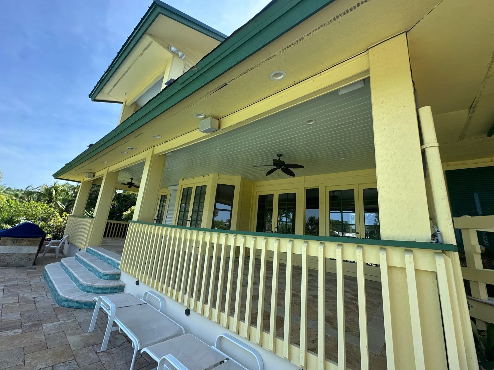 Exterior Painting in Palm City, Florida. Covered porch of a yellow house with pale yellow columns and railings, green trim and a light beadboard ceiling with ceiling fans.