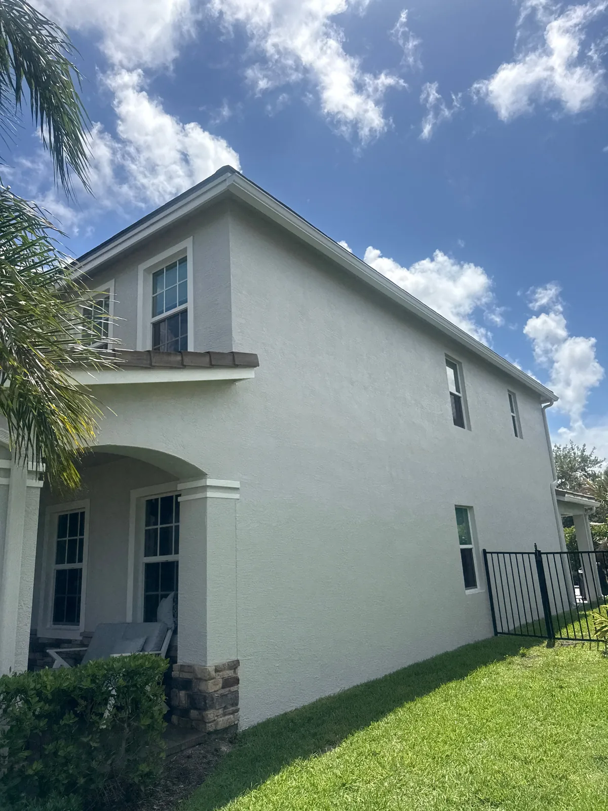 Exterior Painting in Palm City, Florida. Two-storey home exterior in white stucco with a covered porch and stone column bases, green lawn and blue sky with clouds.