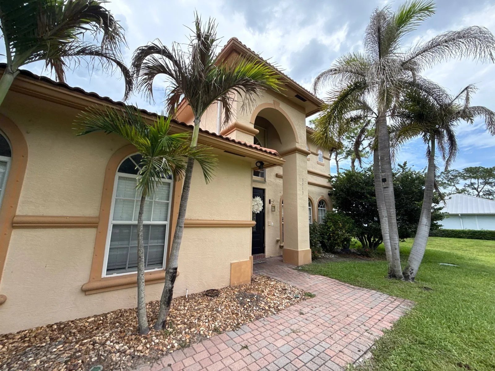 Exterior Painting in Palm City, Florida. Two-storey stucco home painted tan with a barrel-tile roof, arched entry portico, arched window trim and a brick paver walkway.