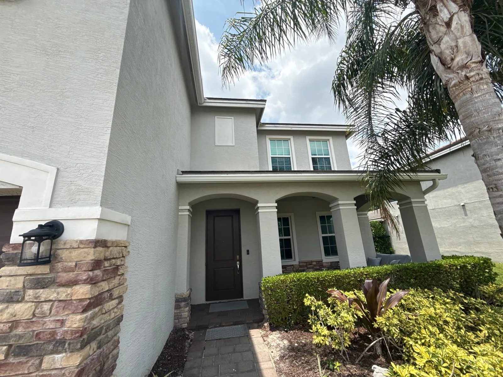Exterior Painting in Palm City, Florida. Two-storey stucco home exterior in grey-green with white trim, covered entry with columns, stone veneer base and dark front door.