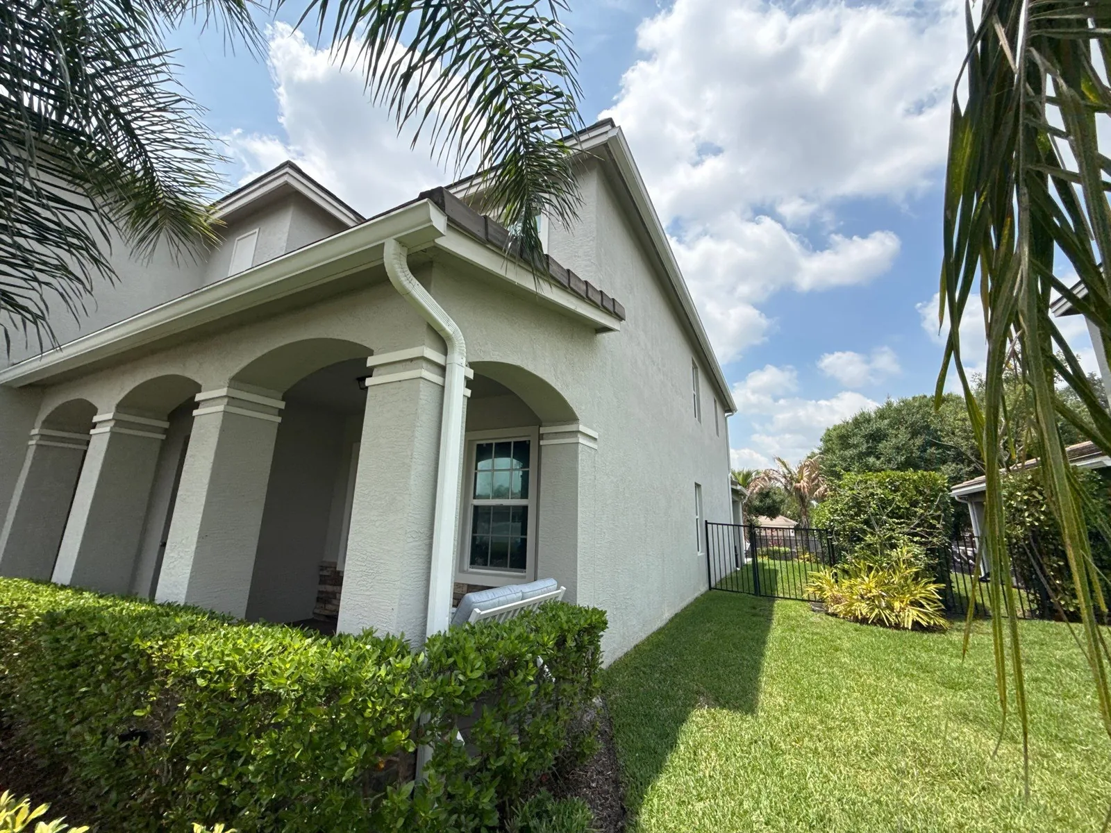Exterior Painting in Palm City, Florida. Two-storey home exterior in light grey stucco with white trim and arched porch columns, hedges and lawn under blue sky.