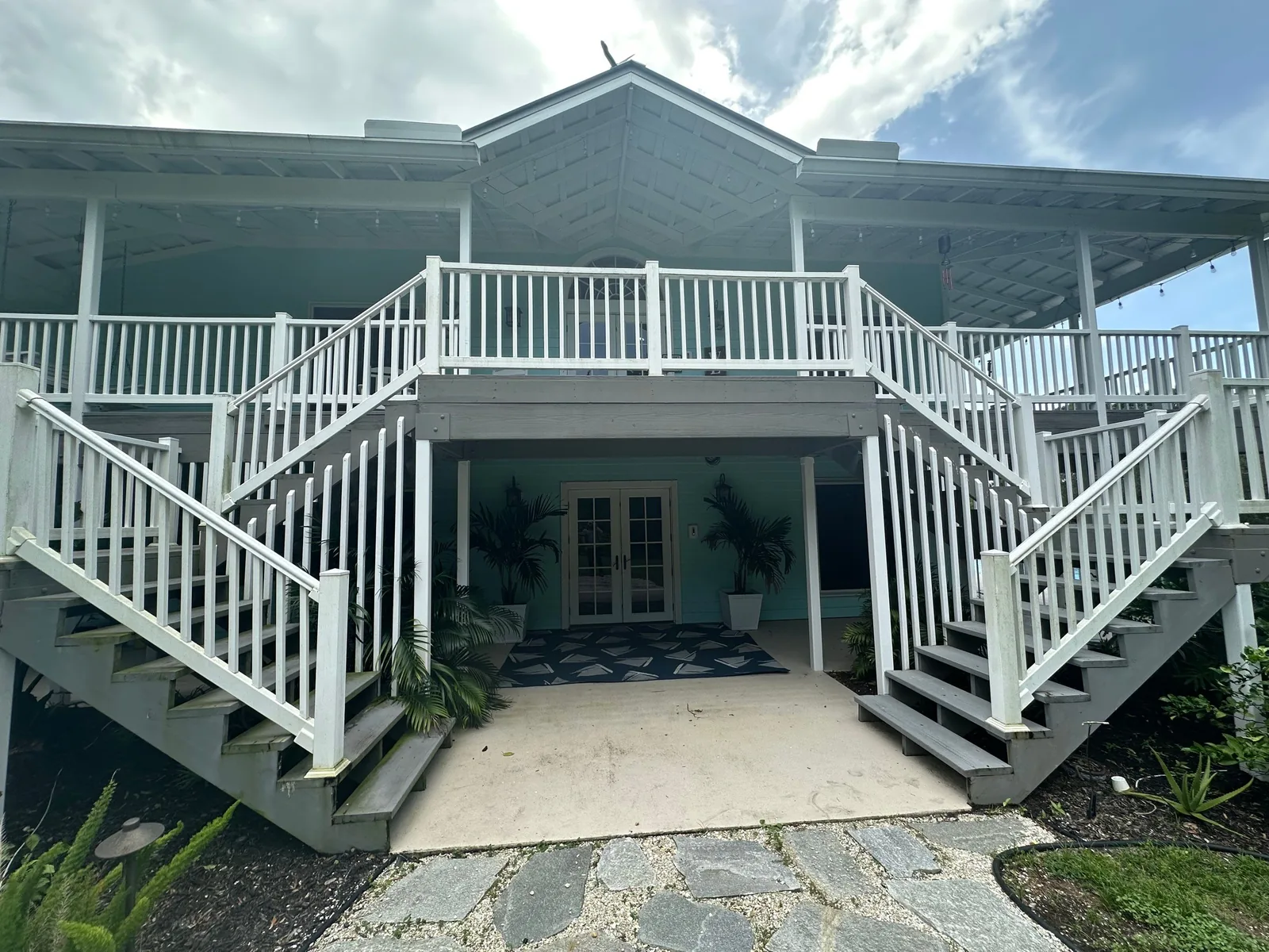 Exterior Painting in Palm City, Florida. Front of a two-storey raised coastal home with green-grey siding, white railings, columns and trim, and twin exterior staircases.