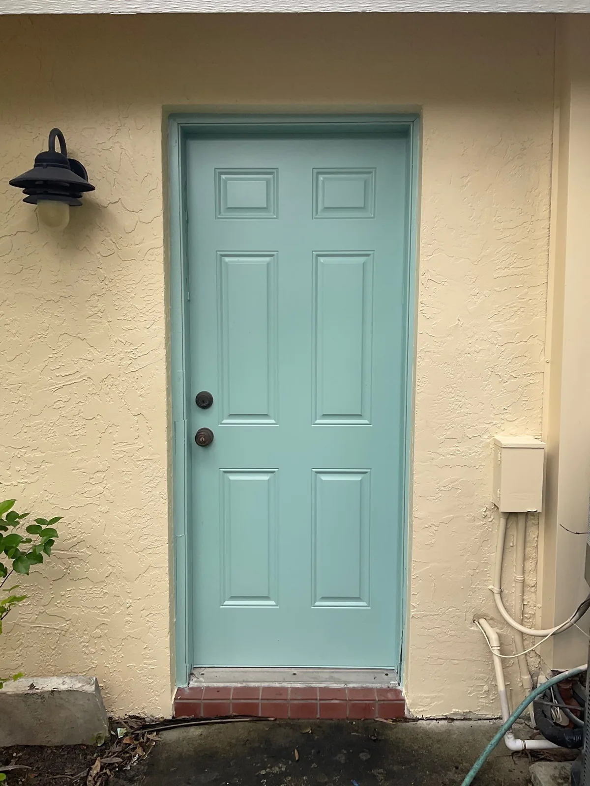Exterior Painting in Palm City, Florida. Exterior six-panel door painted light aqua in a cream stucco wall with a black wall lantern and brick threshold.