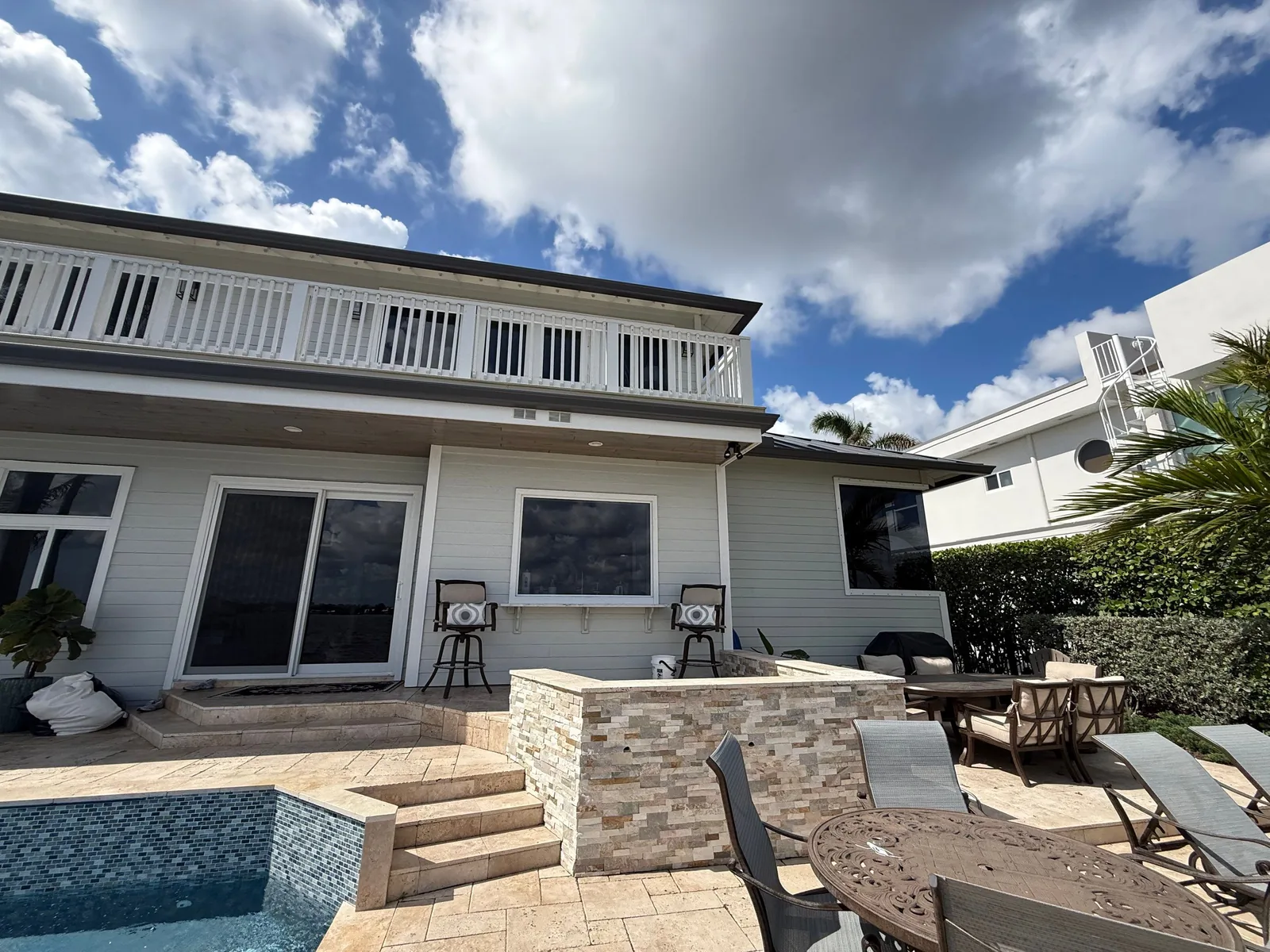 Exterior Painting in Palm City, Florida. Rear elevation of a two-storey home with light grey horizontal siding and white trim, white balcony railing above, pool and paver patio with stone bar in the foreground.