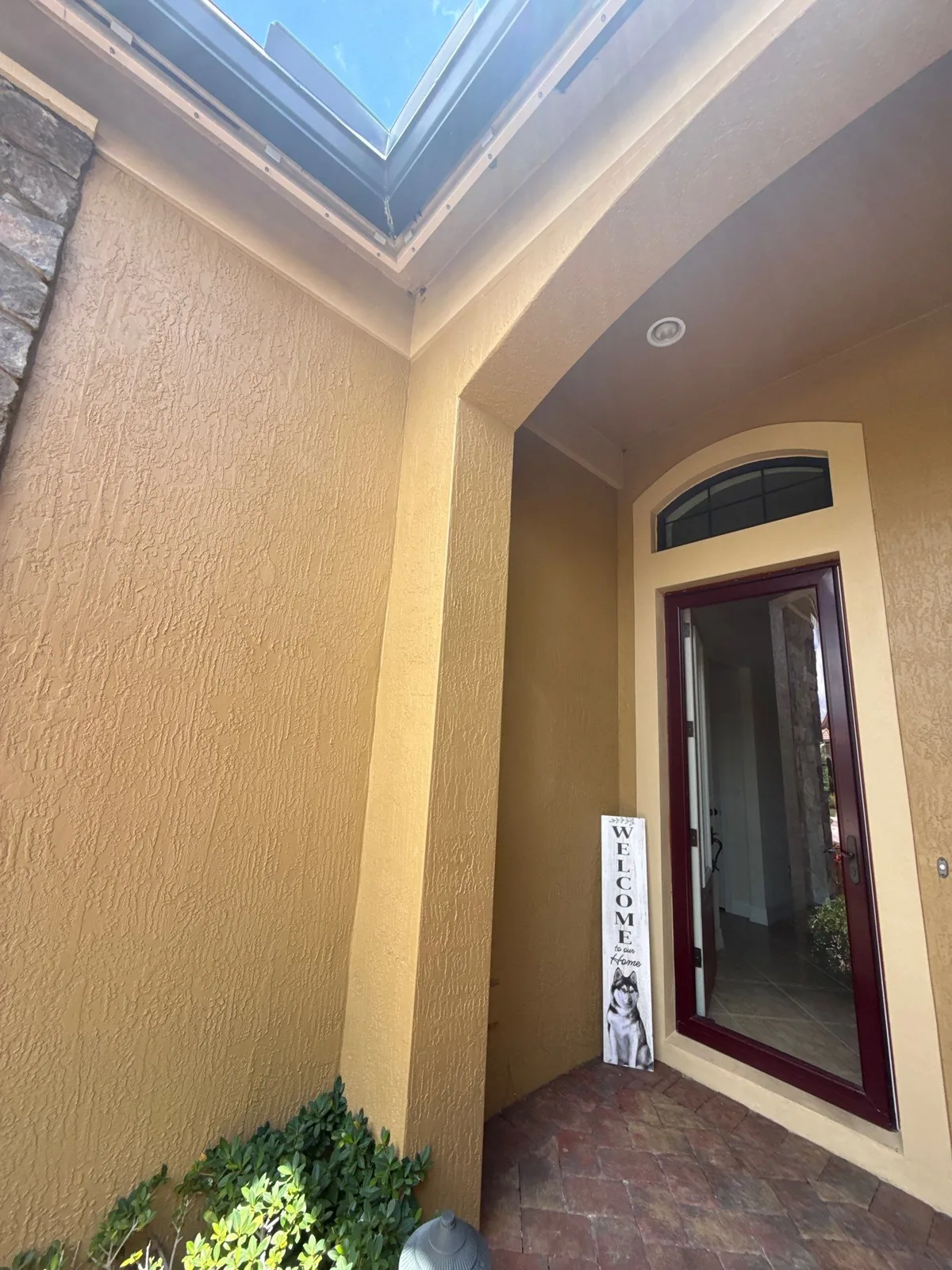 Exterior Painting in Palm City, Florida. Angled view of a home's front entry alcove with gold-toned stucco walls, a dark red glass-panel front door and brick paver flooring.
