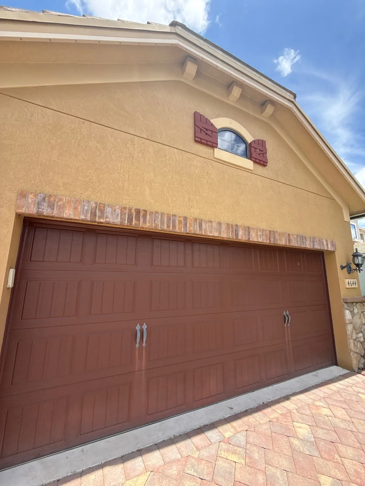 Exterior Painting in Palm City, Florida. Two-car garage with a brown sectional garage door set in gold-toned stucco, decorative shutters and an arched gable window above, brick paver driveway below.