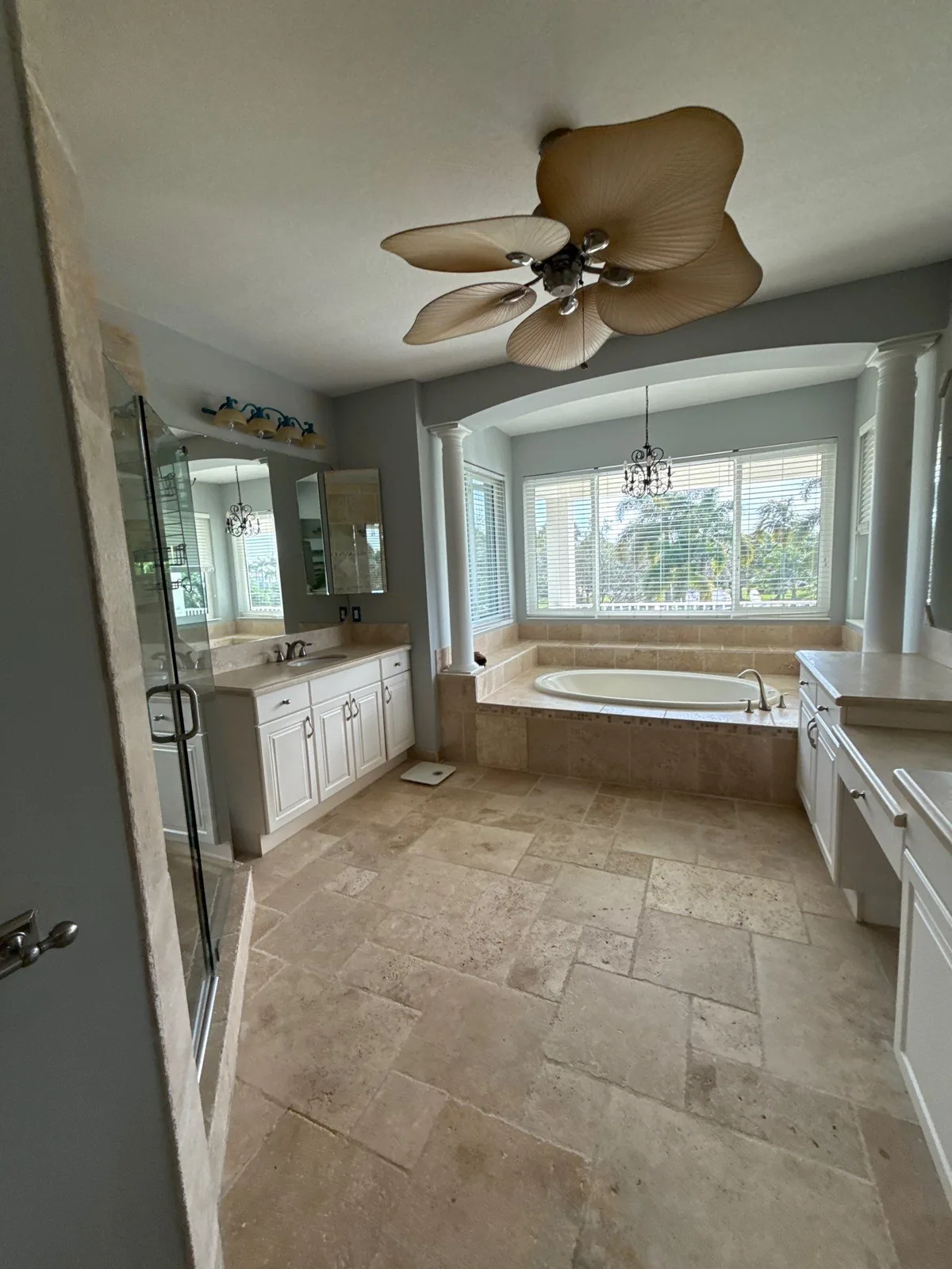 Interior Painting in Palm City, Florida. Master bathroom with grey painted walls, white cabinetry and trim, drop-in tub in a window alcove, glass shower enclosure and travertine tile floor.