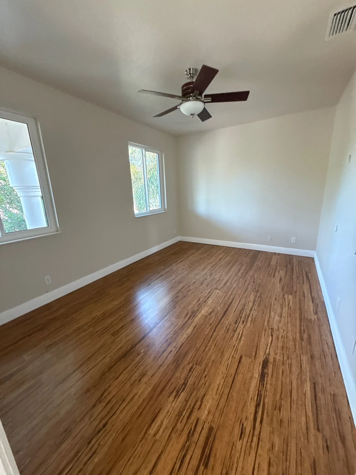 Interior Painting in Palm City, Florida. Empty bedroom with freshly painted light greige walls, white baseboards and window trim, dark wood floor and a ceiling fan.