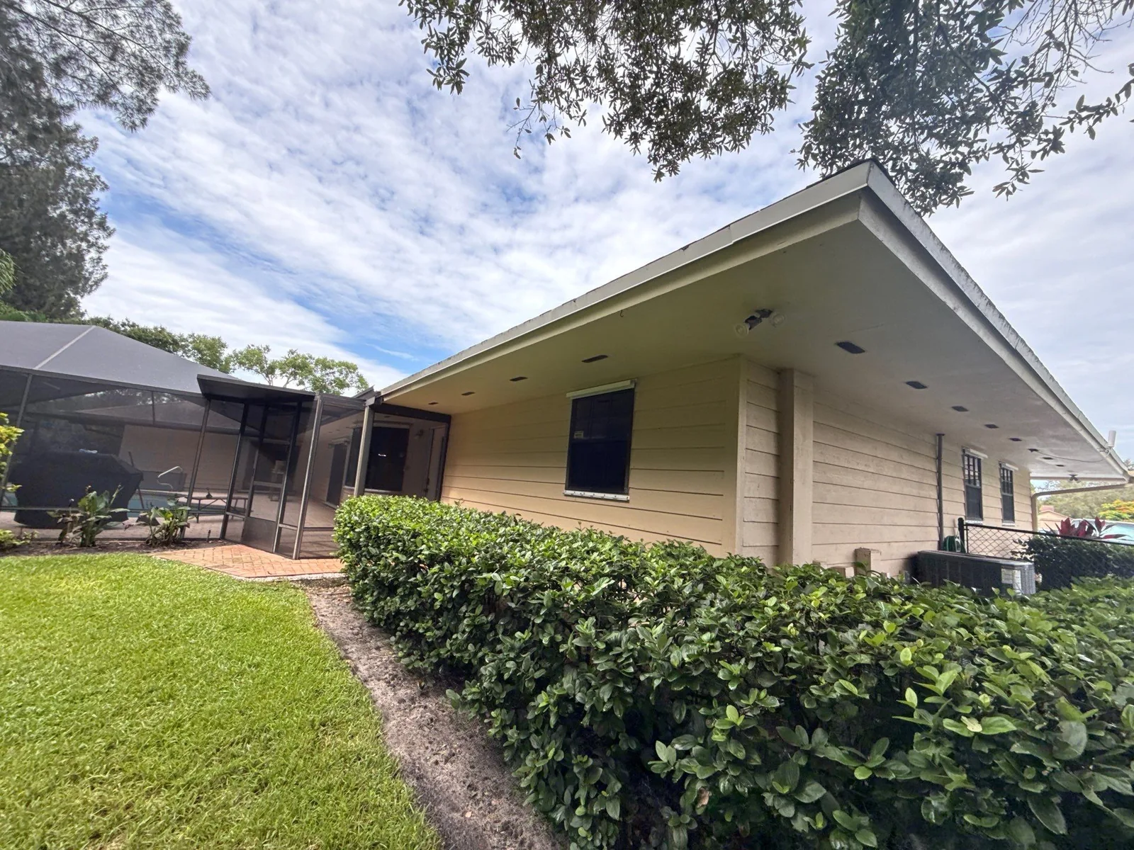 Exterior Painting in Poinciana Park, Florida. Corner of a single-storey home with freshly painted pale yellow horizontal siding, white fascia and soffit, trimmed hedge and lawn, screened enclosure at left.