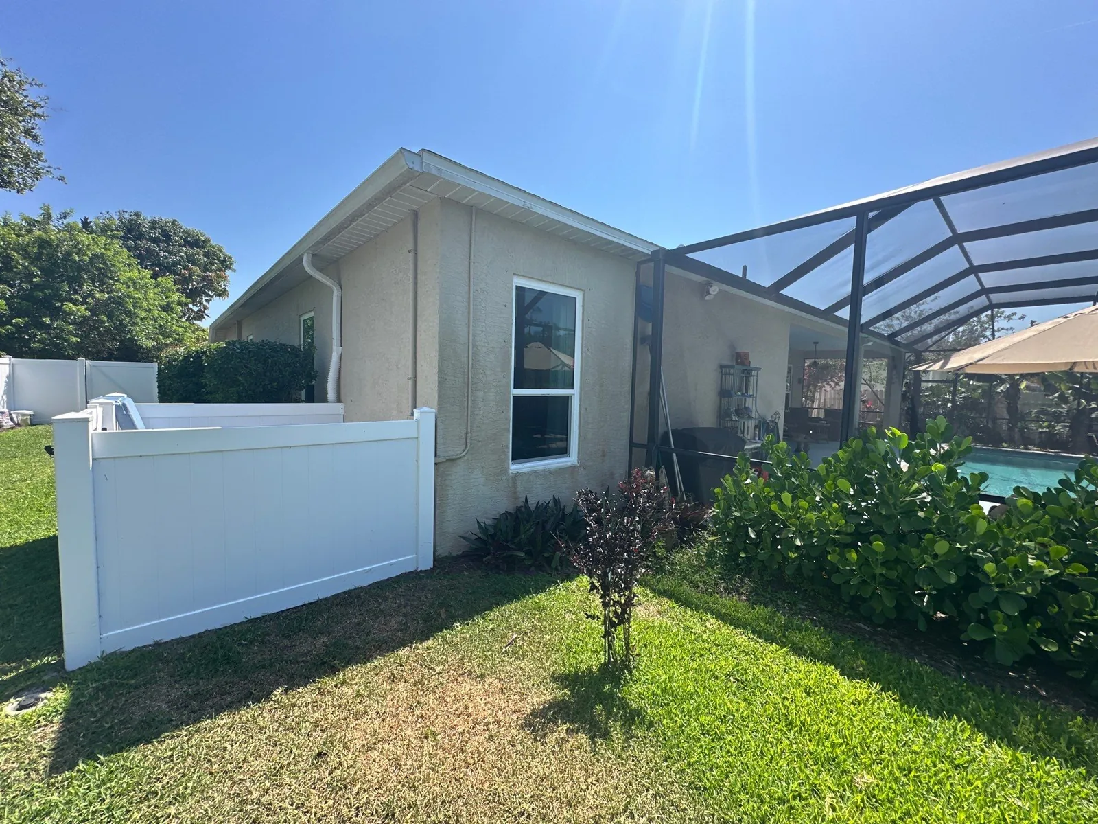 Exterior Painting in Poinciana Park, Florida. Corner elevation of a single-storey stucco home in light beige with white trim, beside a white vinyl fence and a screened pool enclosure.