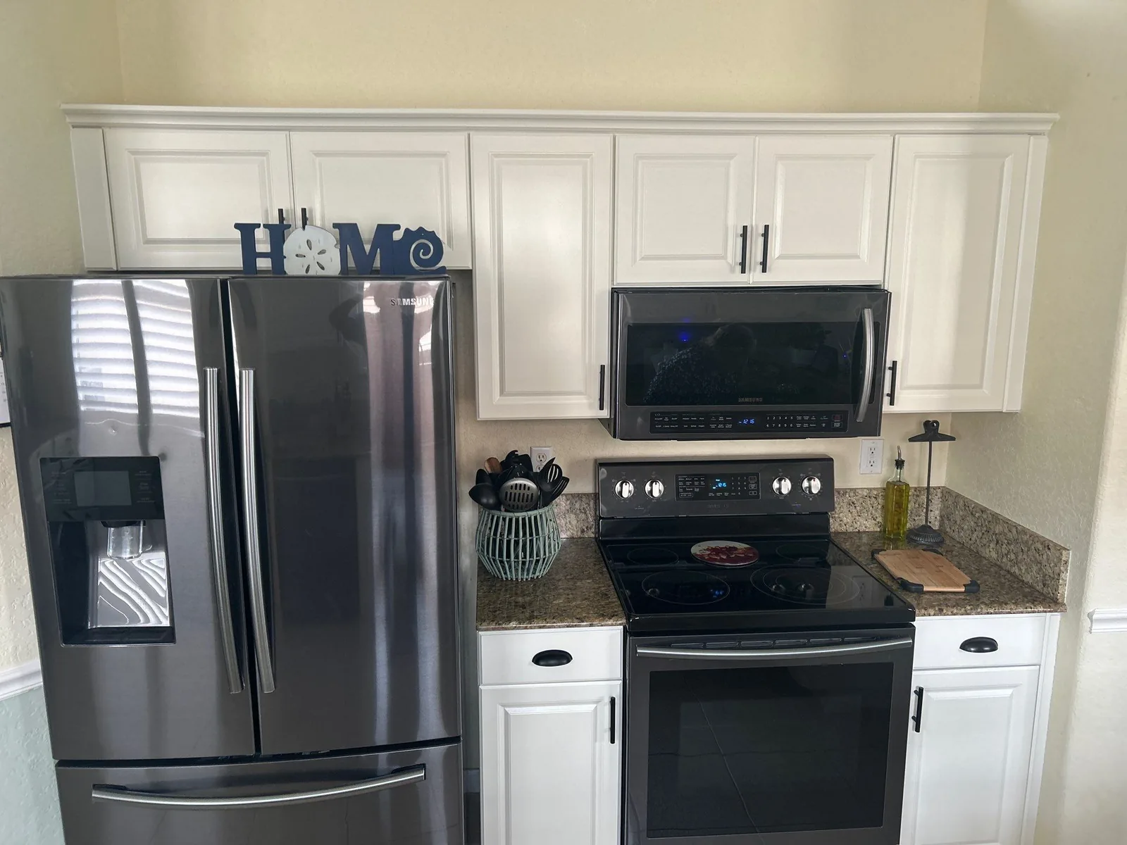 Cabinet Refinishing in Port Saint Lucie, Florida. Kitchen with white painted raised-panel upper and lower cabinets, black range and over-range microwave, stainless refrigerator and speckled granite countertop.