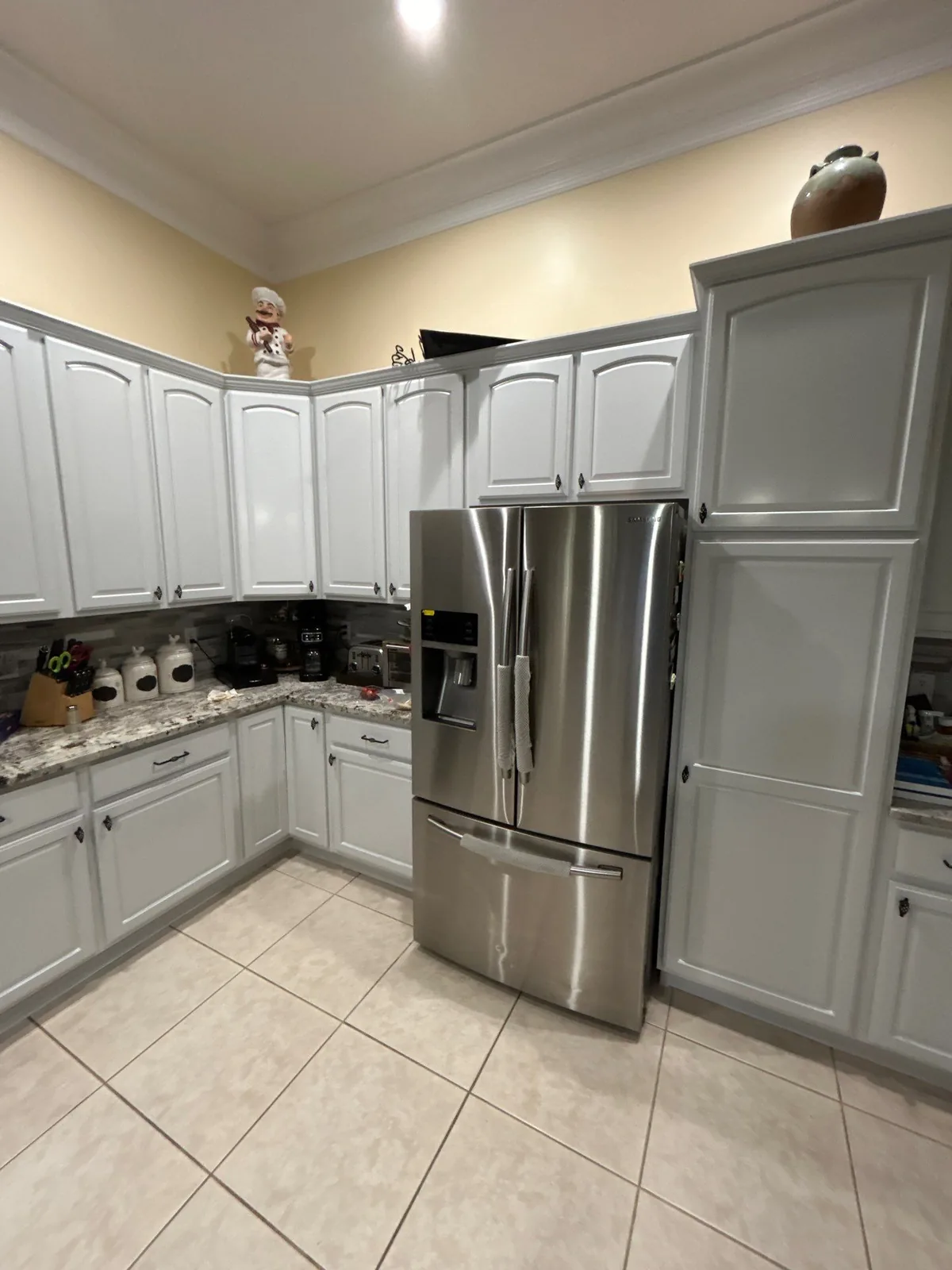 Cabinet Refinishing in Port Saint Lucie, Florida. Refinished kitchen cabinets in light grey with granite countertops, a stainless steel refrigerator and tile flooring.