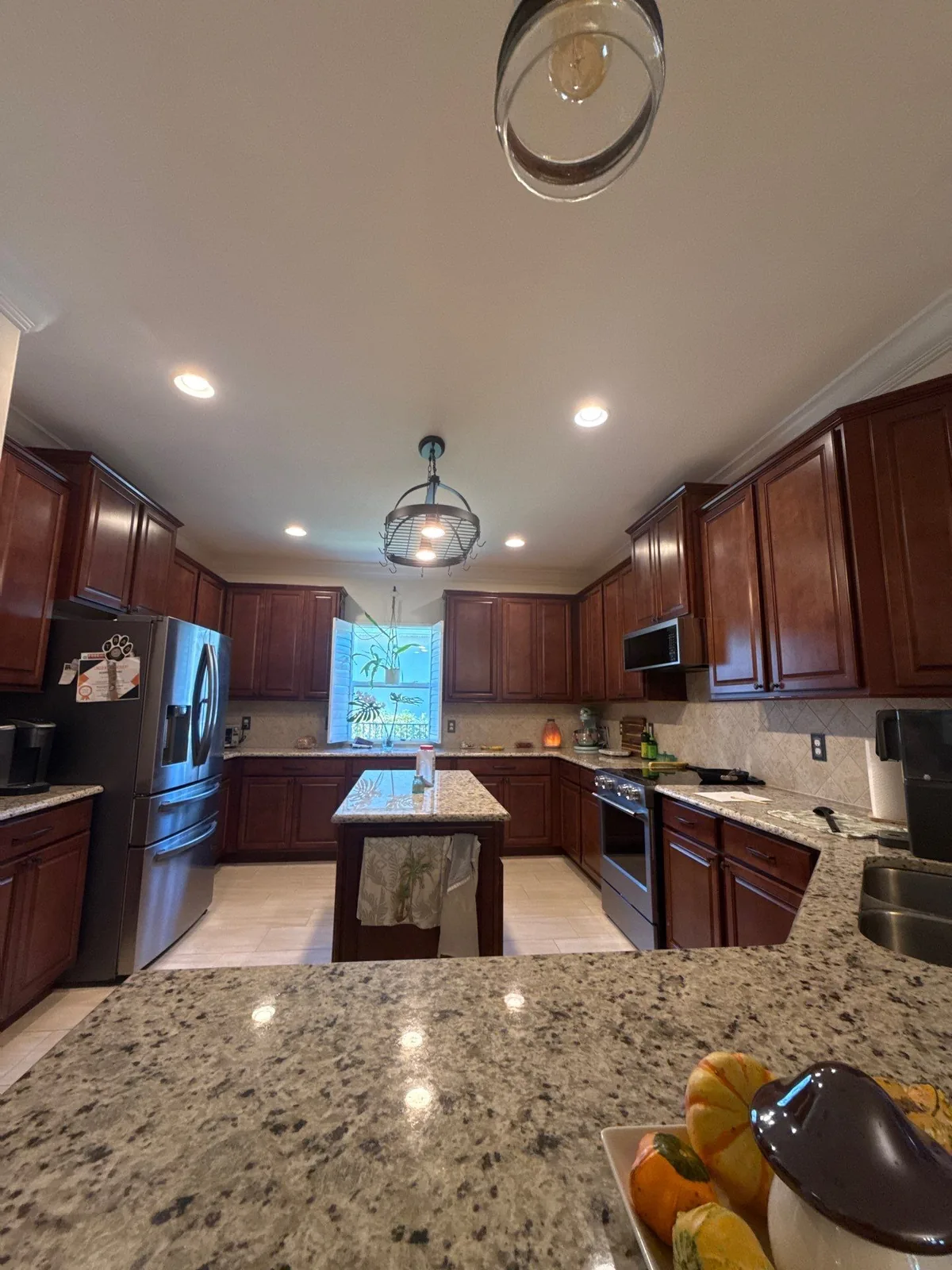 Cabinet Refinishing in Port Saint Lucie, Florida. Kitchen with dark reddish-brown wood cabinets, speckled light granite countertops and centre island, stainless appliances and pendant lights.
