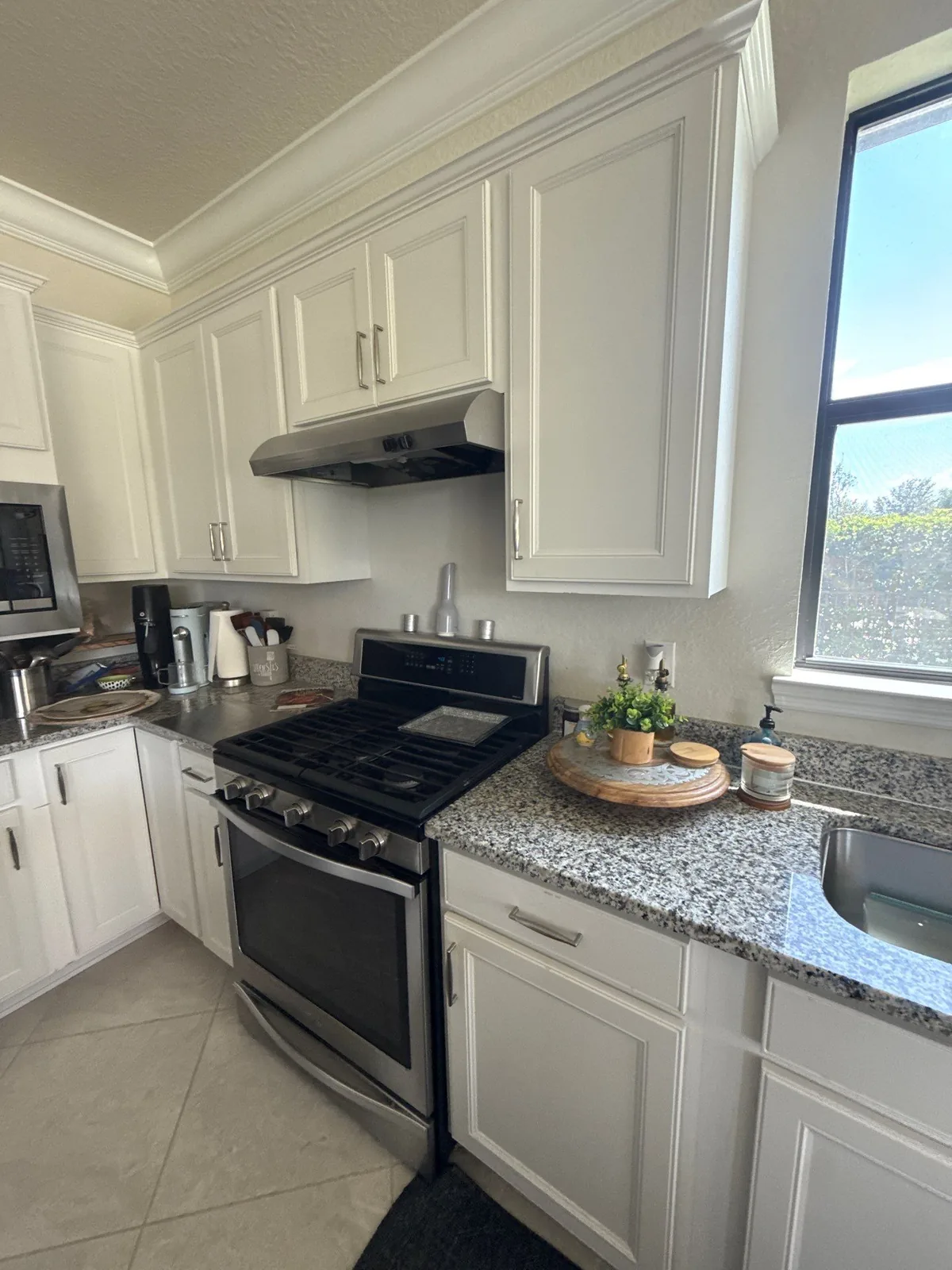 Cabinet Refinishing in Port Saint Lucie, Florida. Kitchen run of white painted shaker cabinets with crown moulding, a wall range hood, stainless gas range and speckled granite countertop, with a bright window at right.