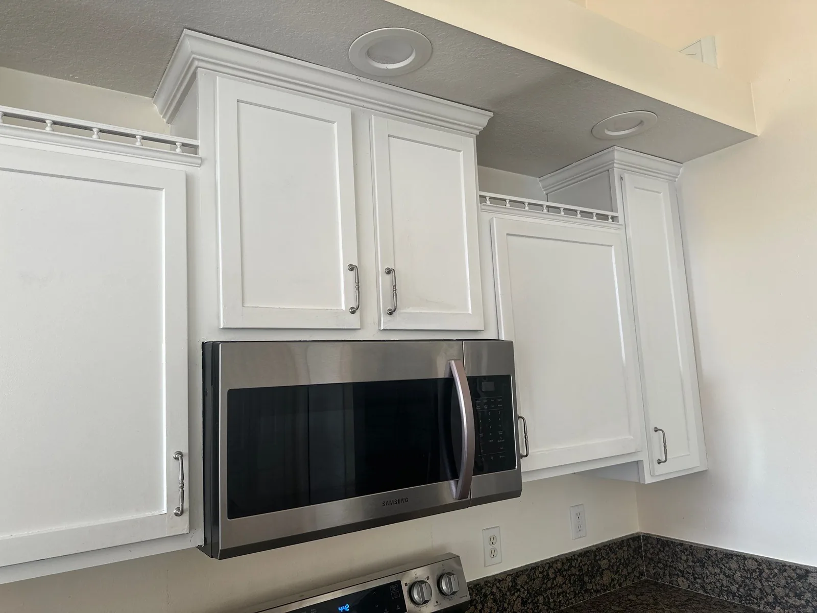 Cabinet Refinishing in Port Saint Lucie, Florida. White-painted upper kitchen cabinets with crown moulding, stainless over-range microwave and dark granite counter.