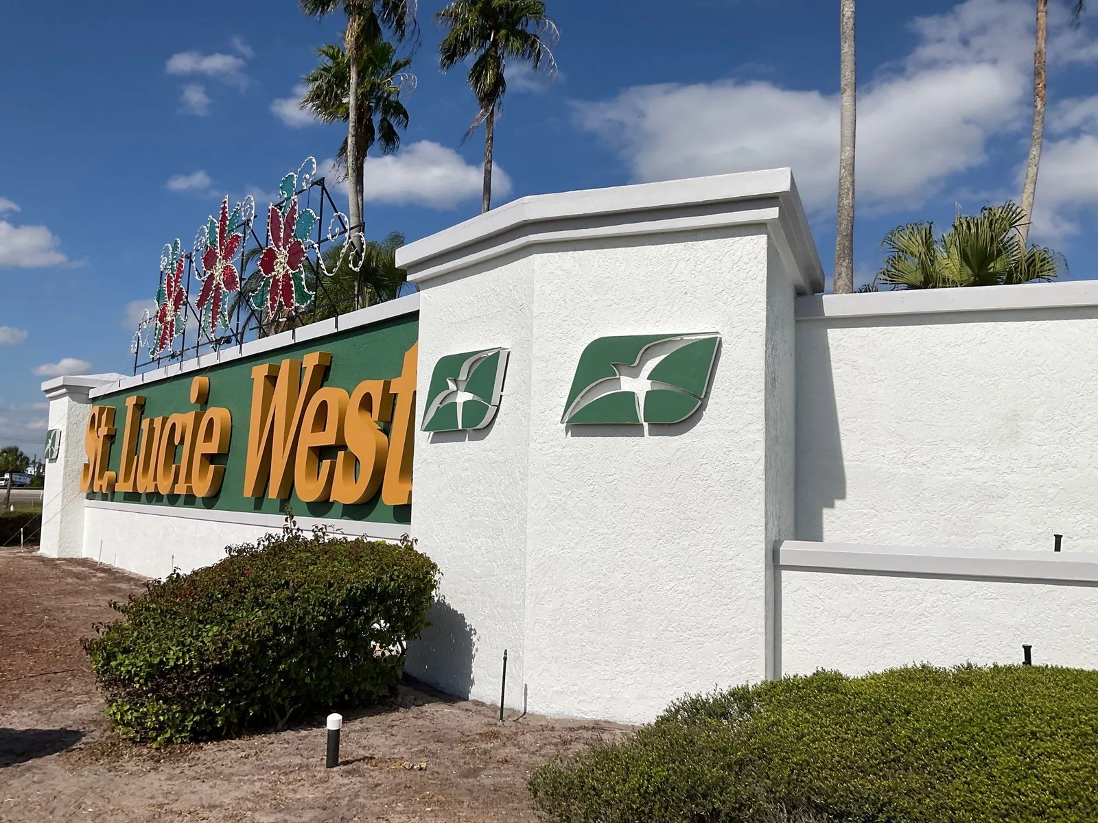 Commercial Painting in Port Saint Lucie, Florida. Freshly painted white stucco community entrance monument wall with raised lettering and green medallion panels, shrubs at the base.