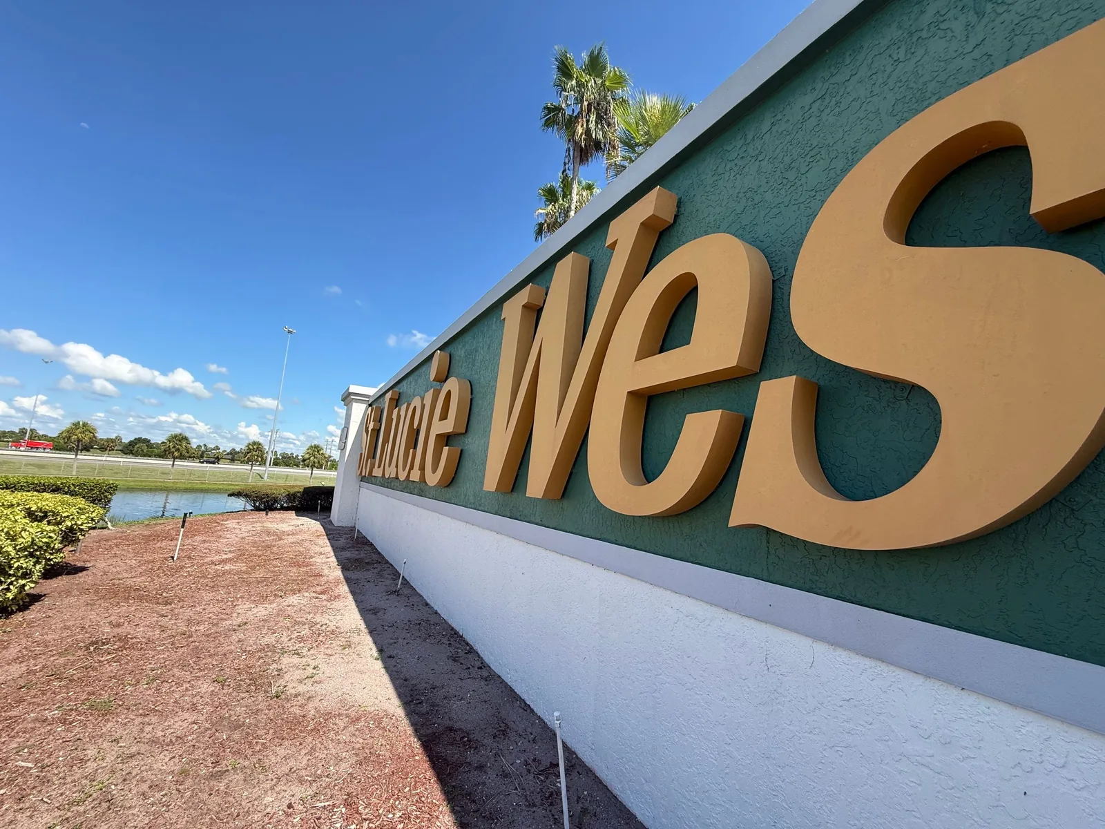 Commercial Painting in Port Saint Lucie, Florida. Community entrance monument sign wall, gold lettering on a green panel above a white painted base, with palms and blue sky.