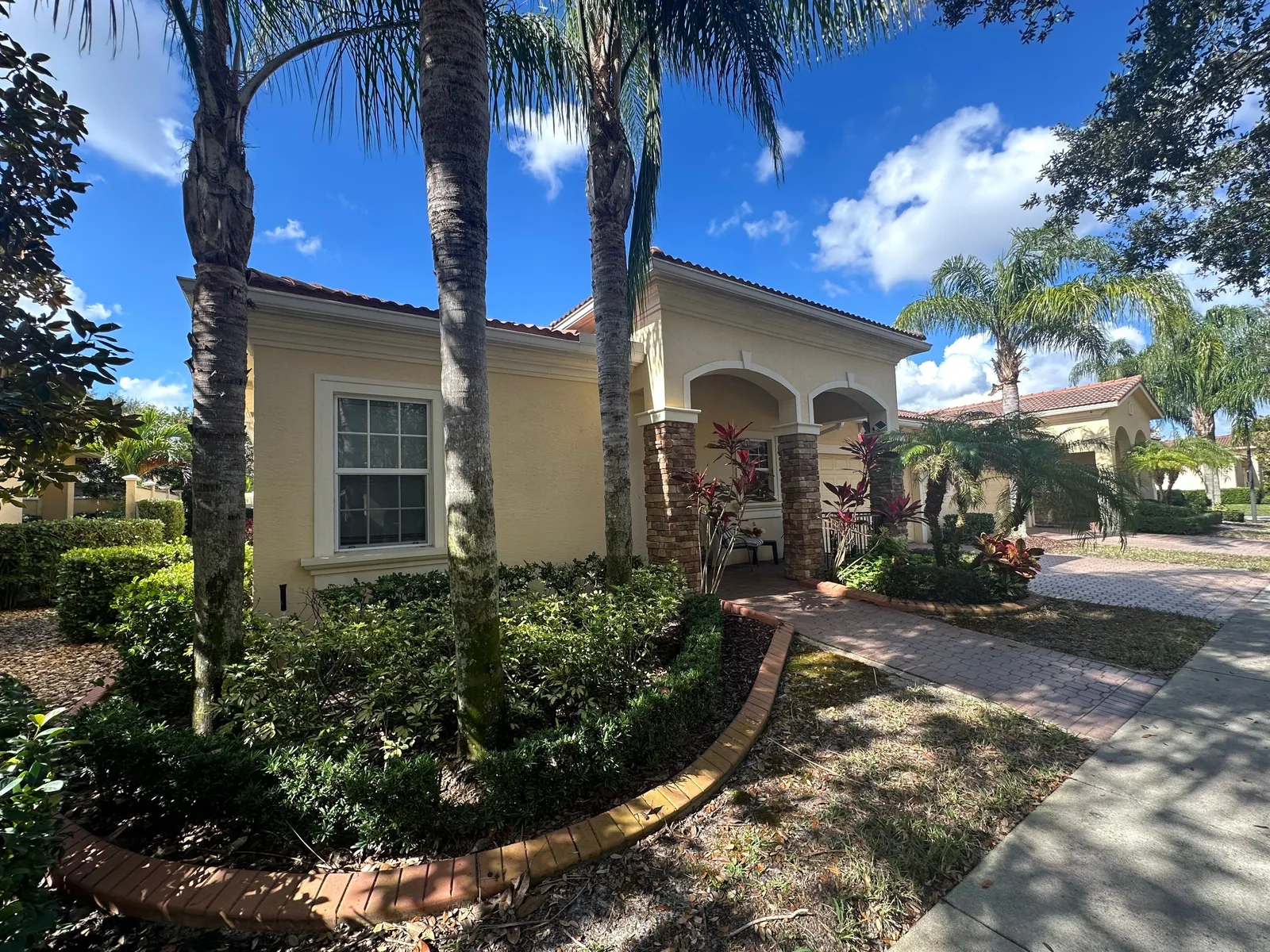 Exterior Painting in Port Saint Lucie, Florida. Single-storey home exterior in pale yellow with an arched entry, tile roof, palms and landscaped walkway.