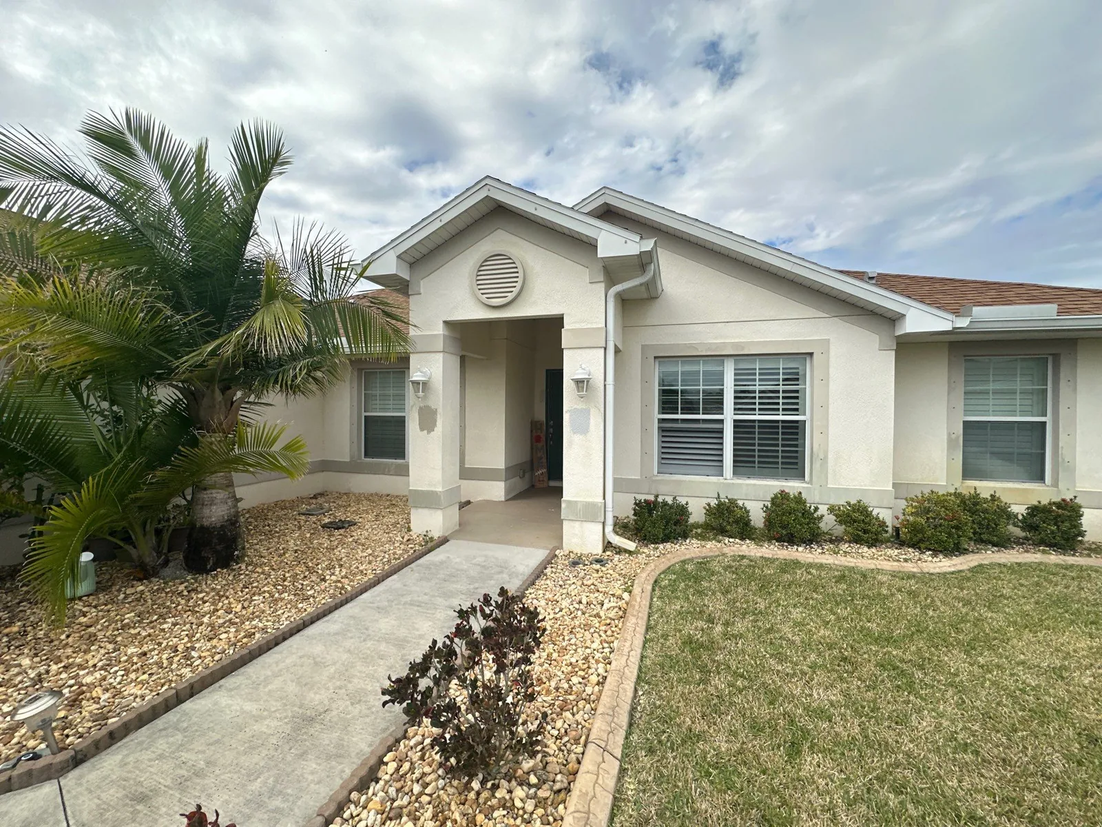 Exterior Painting in Port Saint Lucie, Florida. Front entry of a single-storey stucco home painted cream with white trim, gabled portico with square columns, palm and rock-bed landscaping.