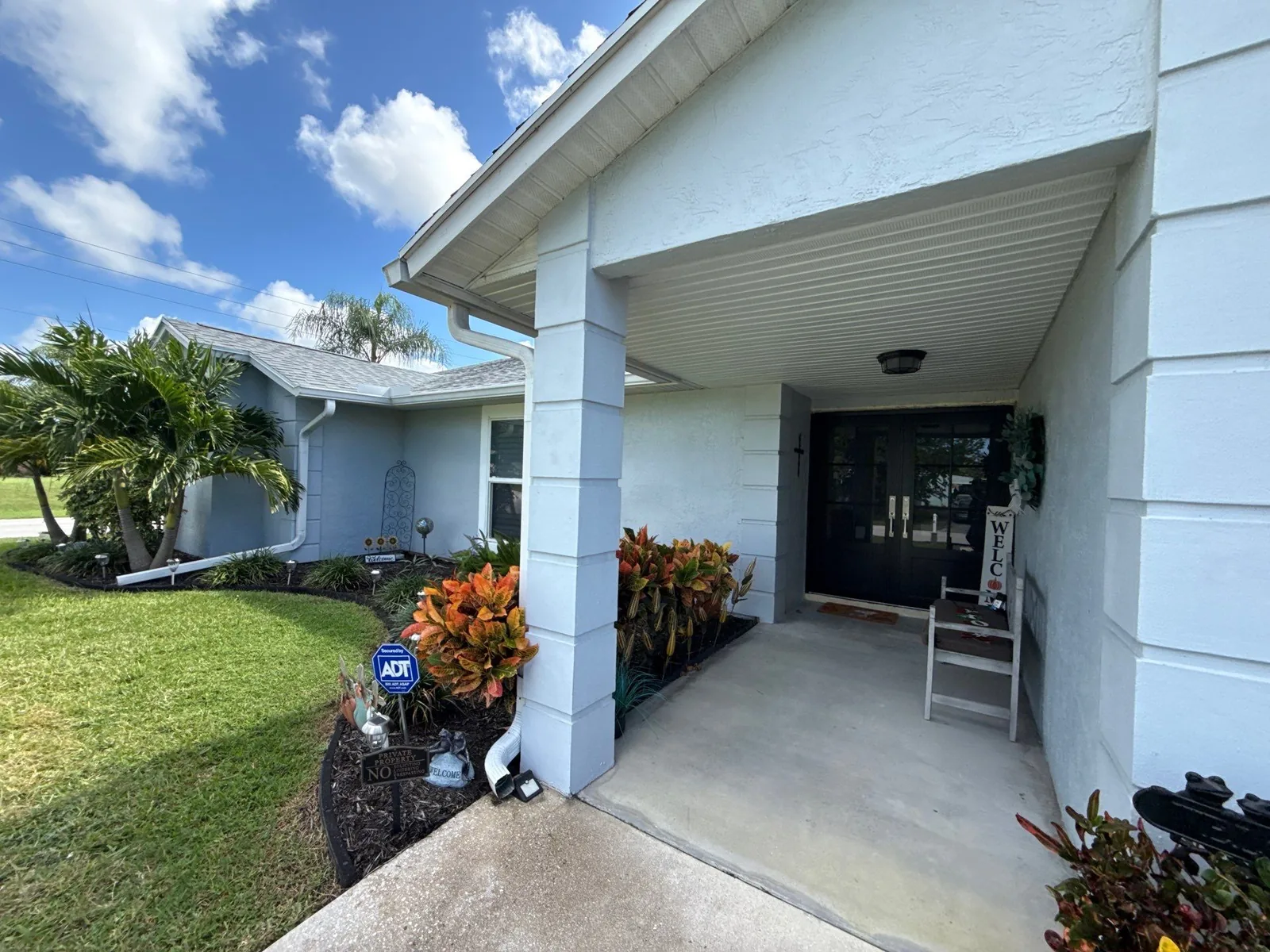 Exterior Painting in Port Saint Lucie, Florida. Single-storey light blue stucco house exterior with white trim and a covered front entry, palm and shrub landscaping.