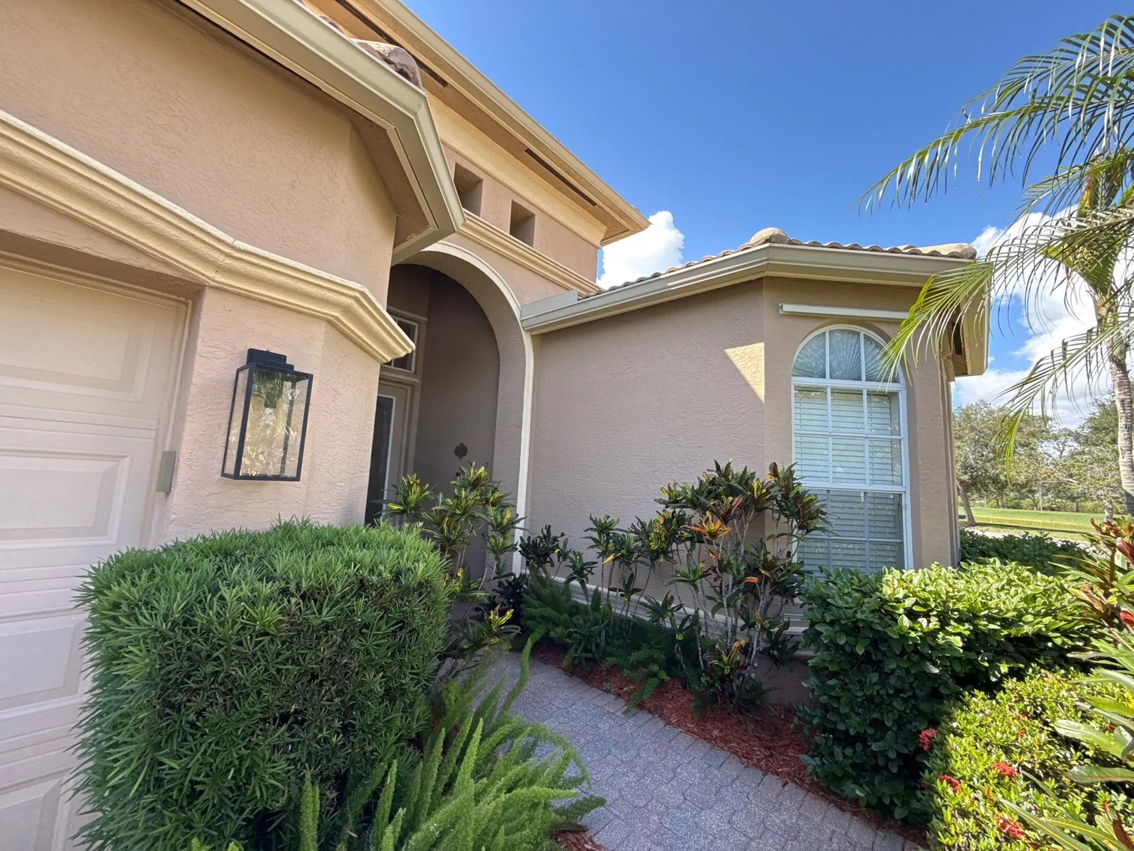 Exterior Painting in Port Saint Lucie, Florida. Single-storey tan stucco home exterior with white window trim, arched entry, tile roof and front landscaping.