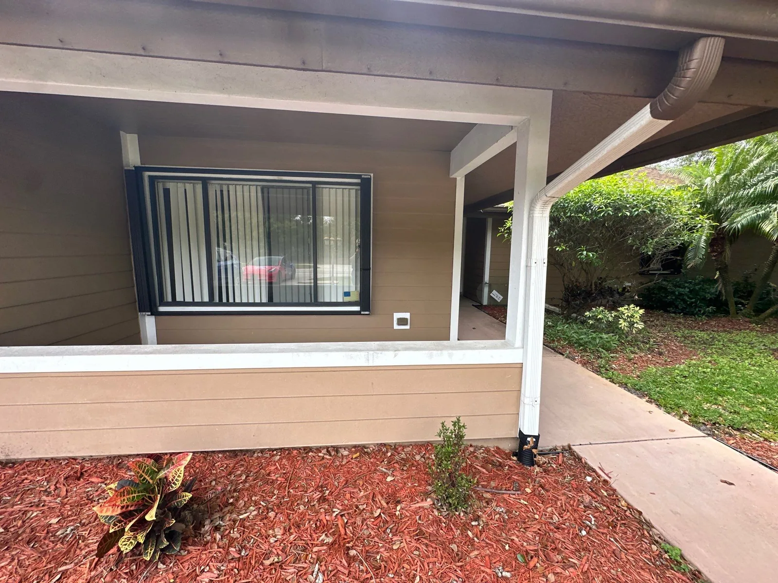 Exterior Painting in Port Saint Lucie, Florida. Covered front porch of a single-storey home with brown horizontal siding, white trim, posts and railing, and a mulched planting bed.