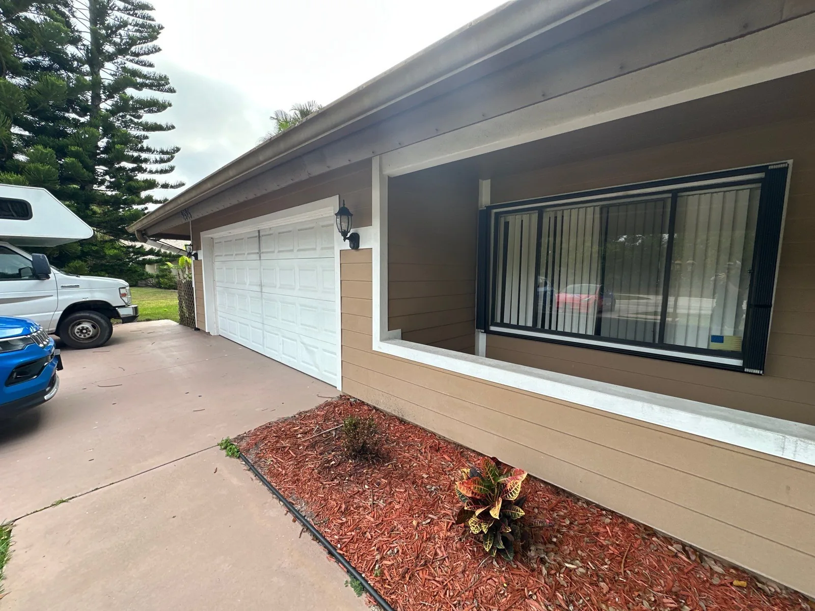 Exterior Painting in Port Saint Lucie, Florida. Front of a single-storey home with brown siding, white trim, white garage door and a mulched planting bed.