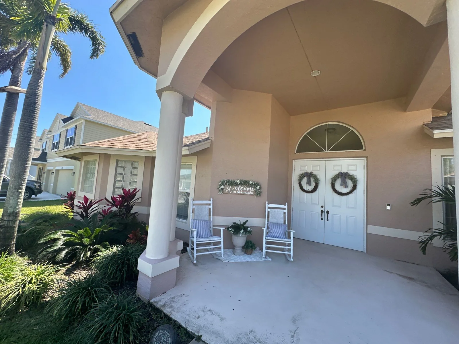 Exterior Painting in Port Saint Lucie, Florida. Covered front entry of a house with pink stucco walls, white columns and trim, and white double front doors with wreaths.