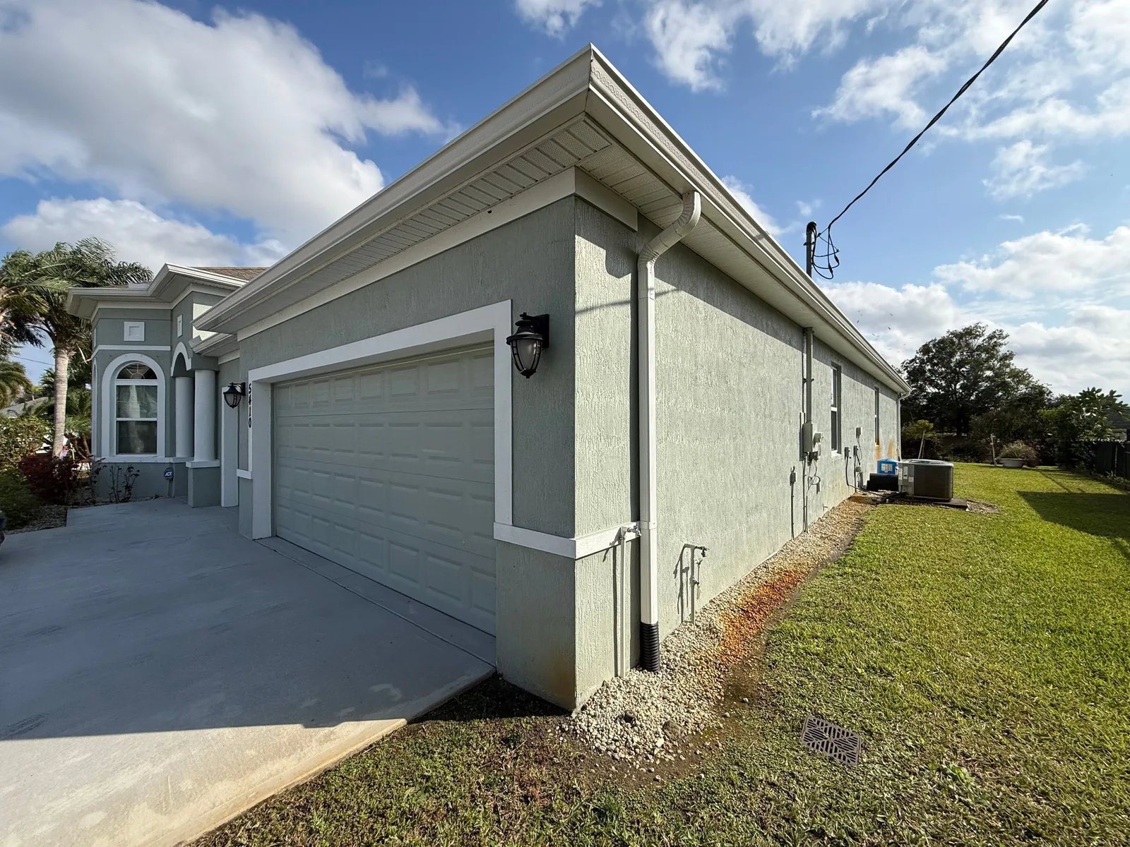 Exterior Painting in Port Saint Lucie, Florida. Corner view of a single-storey stucco house painted pale sage green with white trim and fascia, a white two-car garage door, concrete driveway and lawn in full sun.