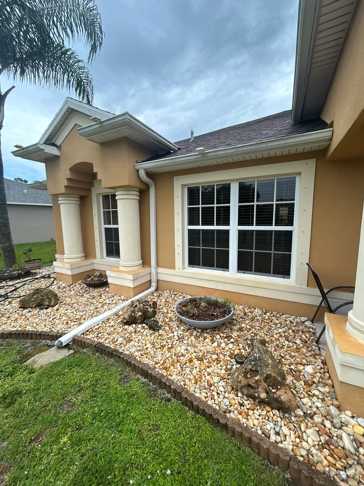 Exterior Painting in Port Saint Lucie, Florida. Front elevation of a tan stucco house with white trim, a white-framed bay window, arched entry and round columns above a river-rock bed.