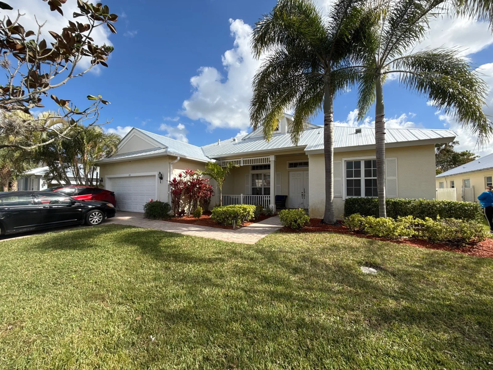 Exterior Painting in Port Saint Lucie, Florida. Single-storey house front elevation in off-white with white trim and metal roof, palm trees, lawn and cars in the driveway.