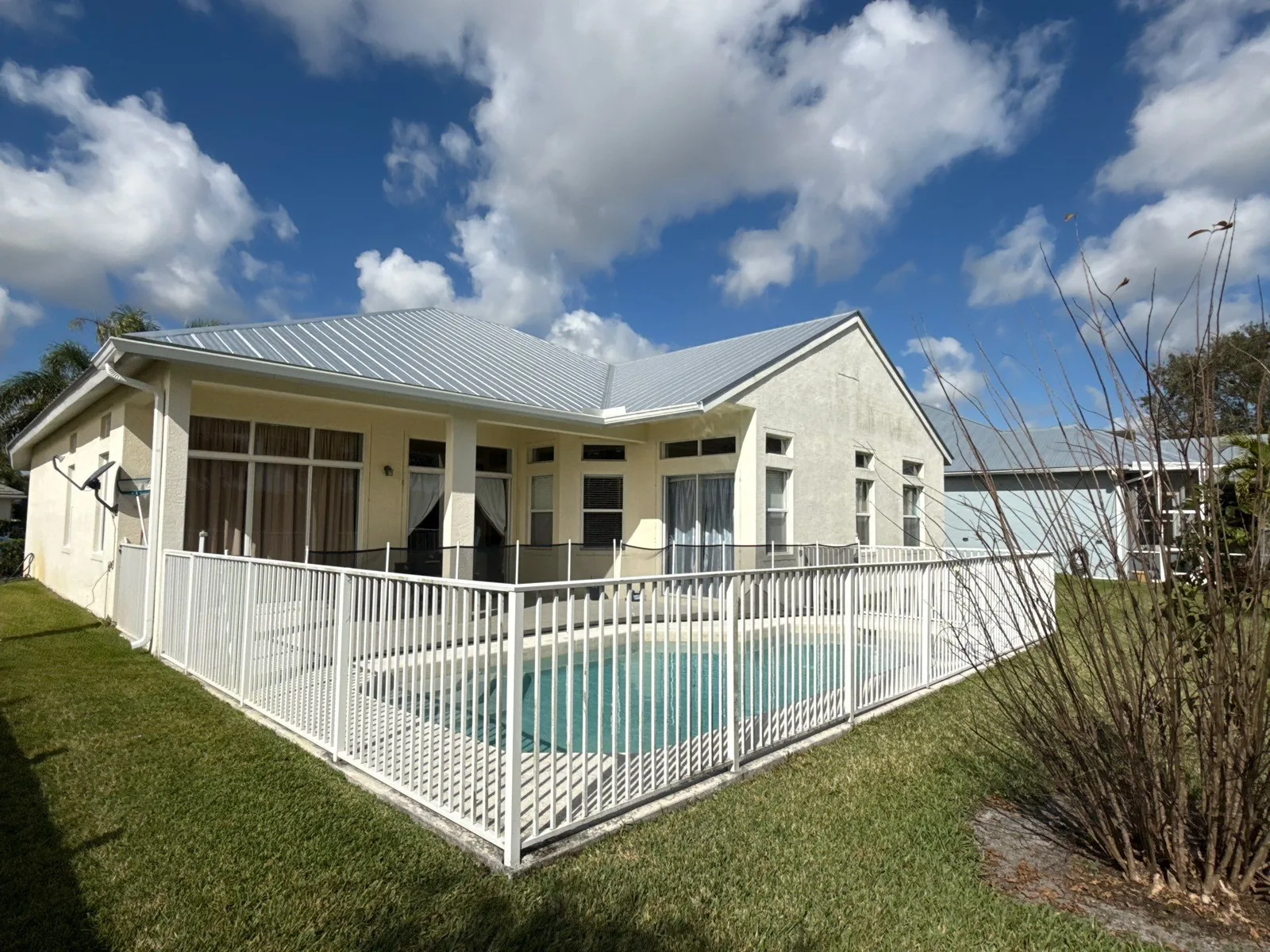 Exterior Painting in Port Saint Lucie, Florida. Rear elevation of a single-storey off-white house with a metal roof, screened porch and a fenced pool in the foreground.