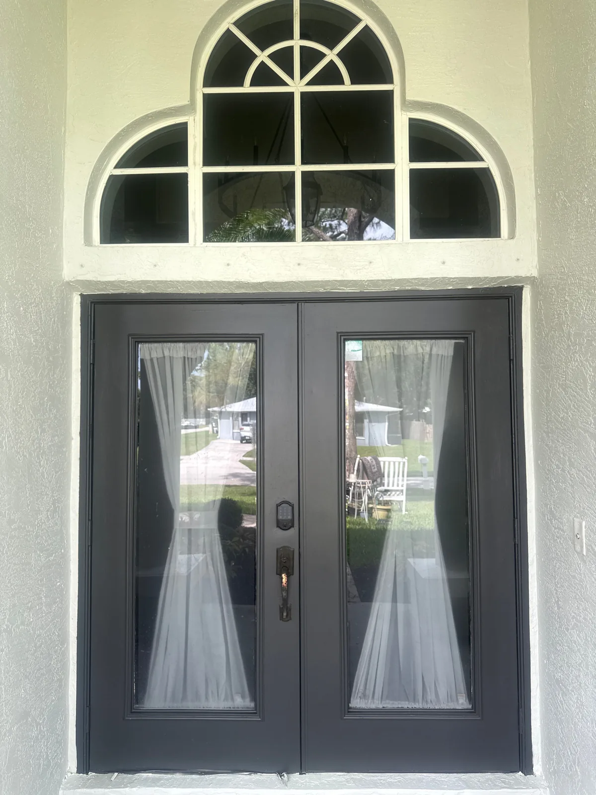 Exterior Painting in Port Saint Lucie, Florida. Pair of dark-painted glass front doors with sheer curtains, set in a cream stucco surround beneath an arched fanlight window.
