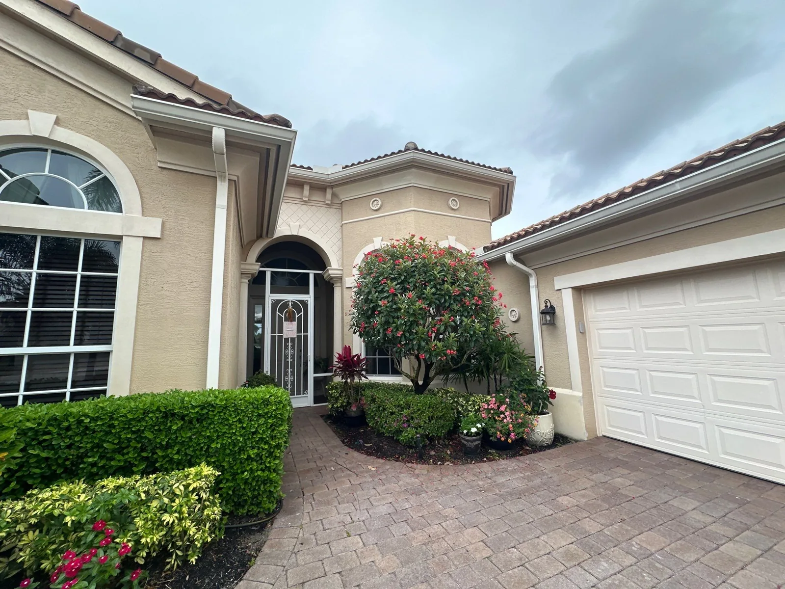 Exterior Painting in Port Saint Lucie, Florida. Front elevation of a single-storey home with cream stucco walls, white trim, arched entry with a tile roof, and a paver driveway.