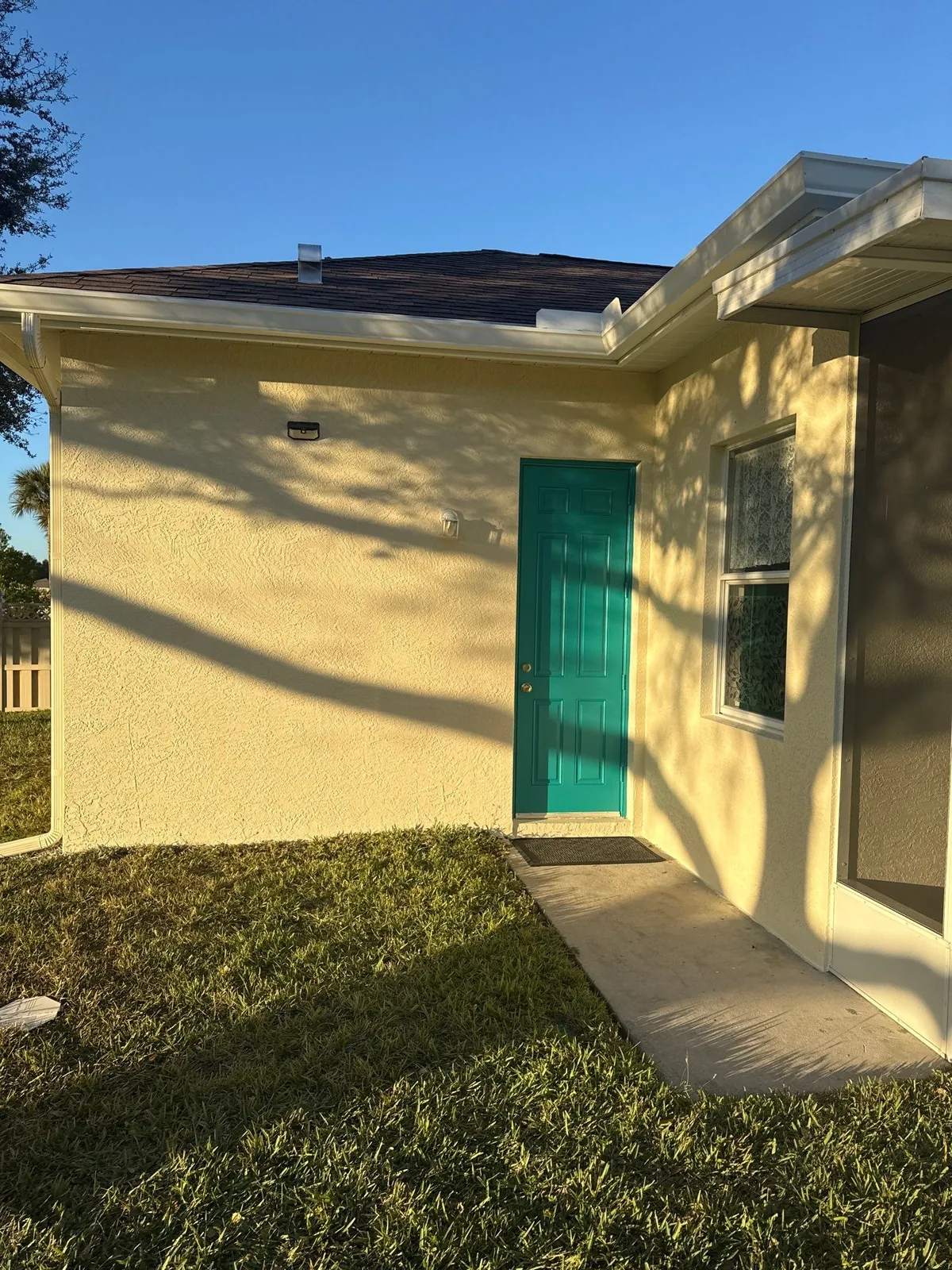 Exterior Painting in Port Saint Lucie, Florida. Corner of a single-storey house painted pale yellow with a teal side door and white window trim.
