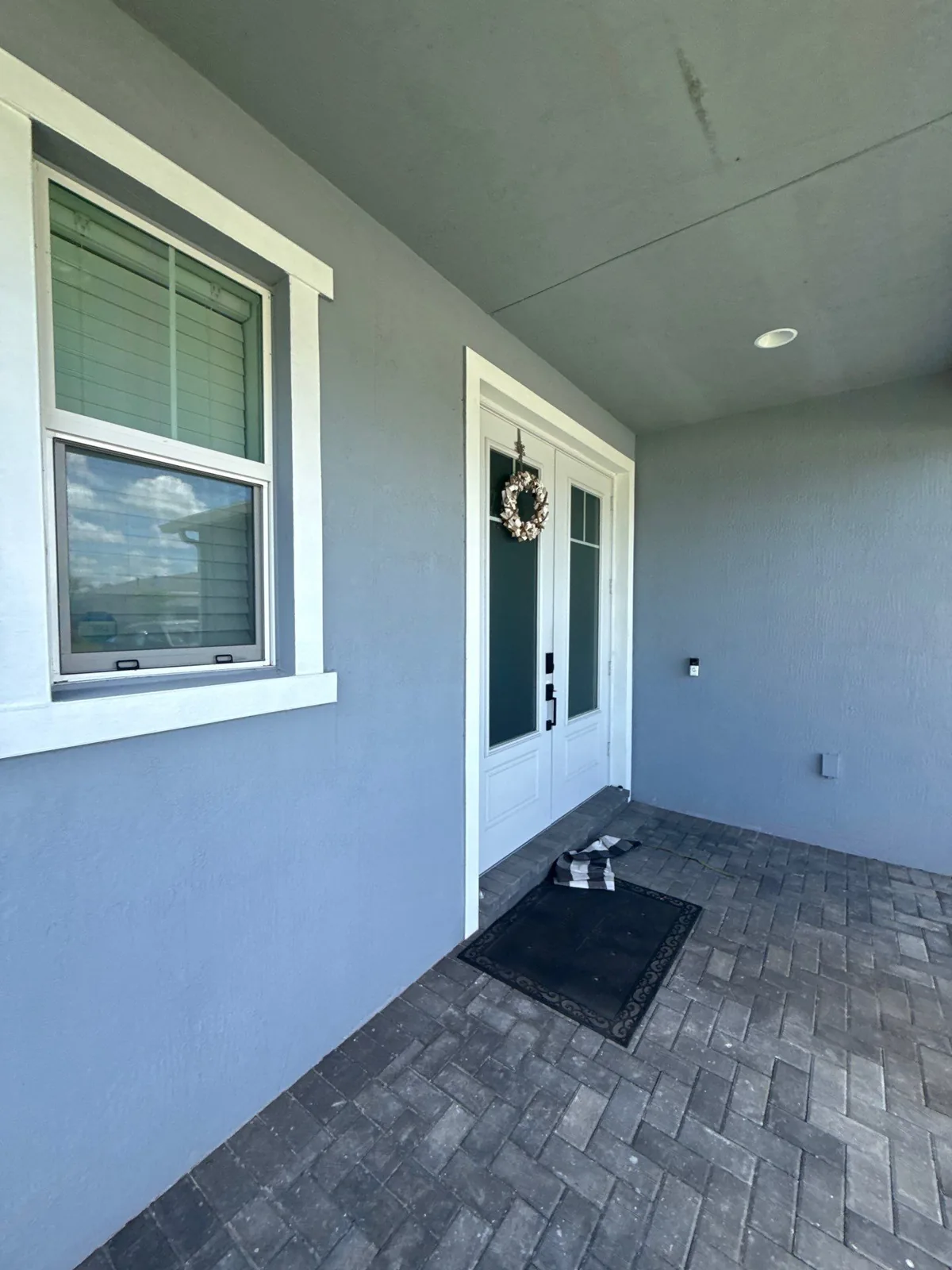Exterior Painting in Port Saint Lucie, Florida. Covered front entry with blue-grey stucco walls, white trim, white double doors with a wreath and a paver porch.