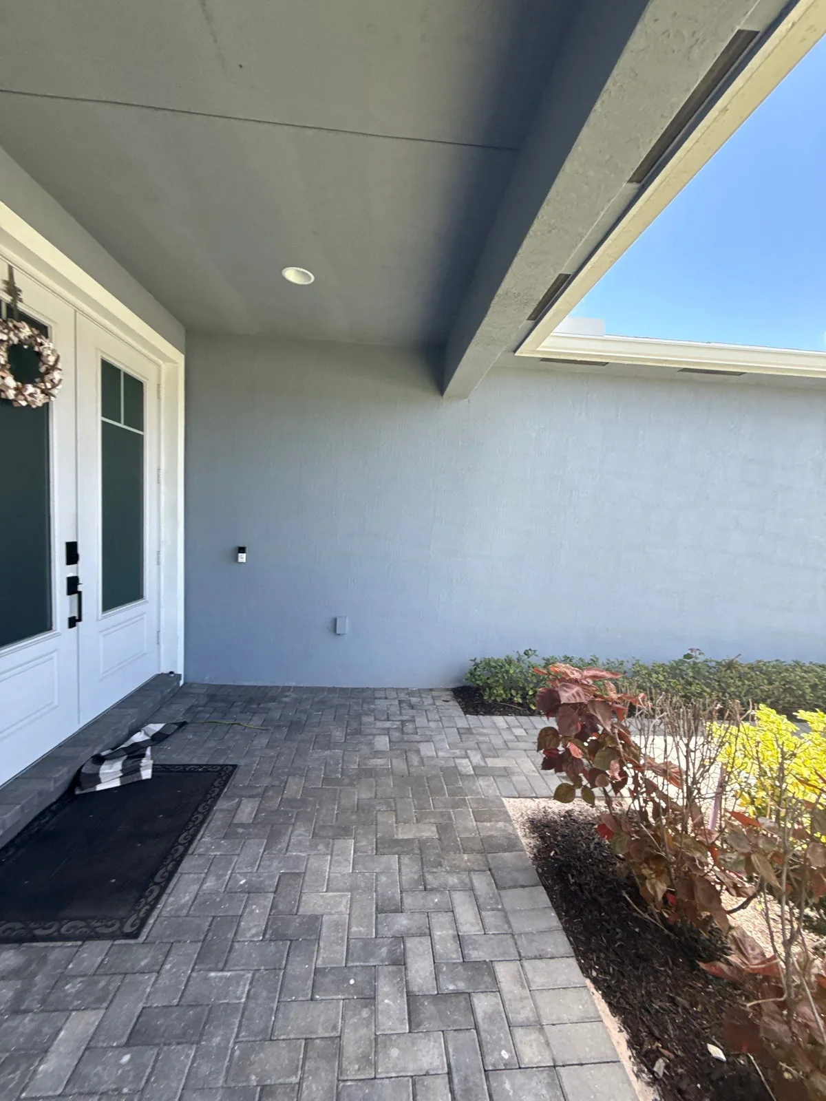 Exterior Painting in Port Saint Lucie, Florida. Front entry courtyard with grey stucco walls, white door and sidelight, paver paving and planted beds.