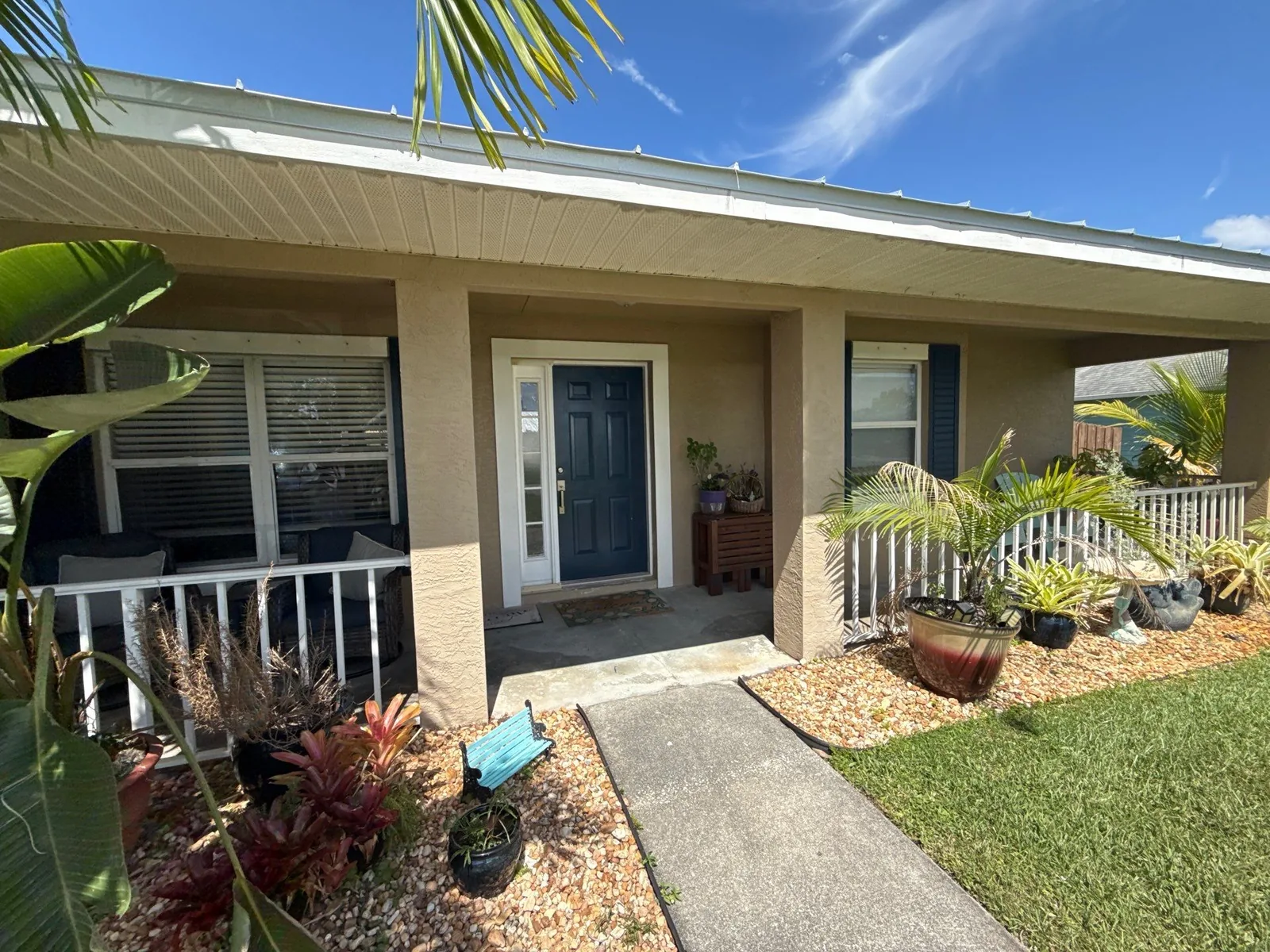 Exterior Painting in Port Saint Lucie, Florida. Covered front entry of a tan stucco home with a dark teal door and shutters, white porch railing and potted plants.