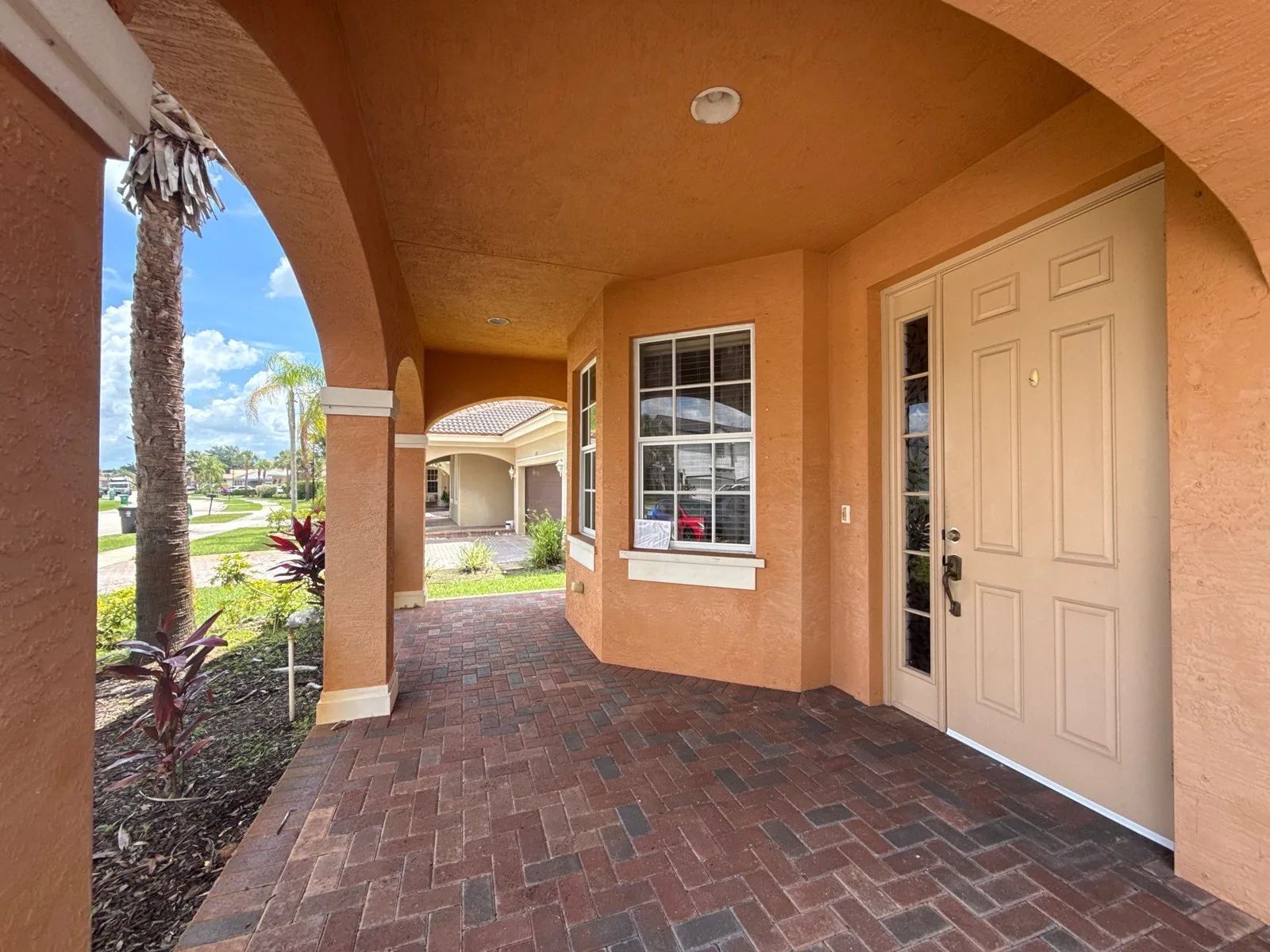 Exterior Painting in Port Saint Lucie, Florida. Covered front entry of a stucco home with salmon walls, arched columns, white front door and brick paver walkway.