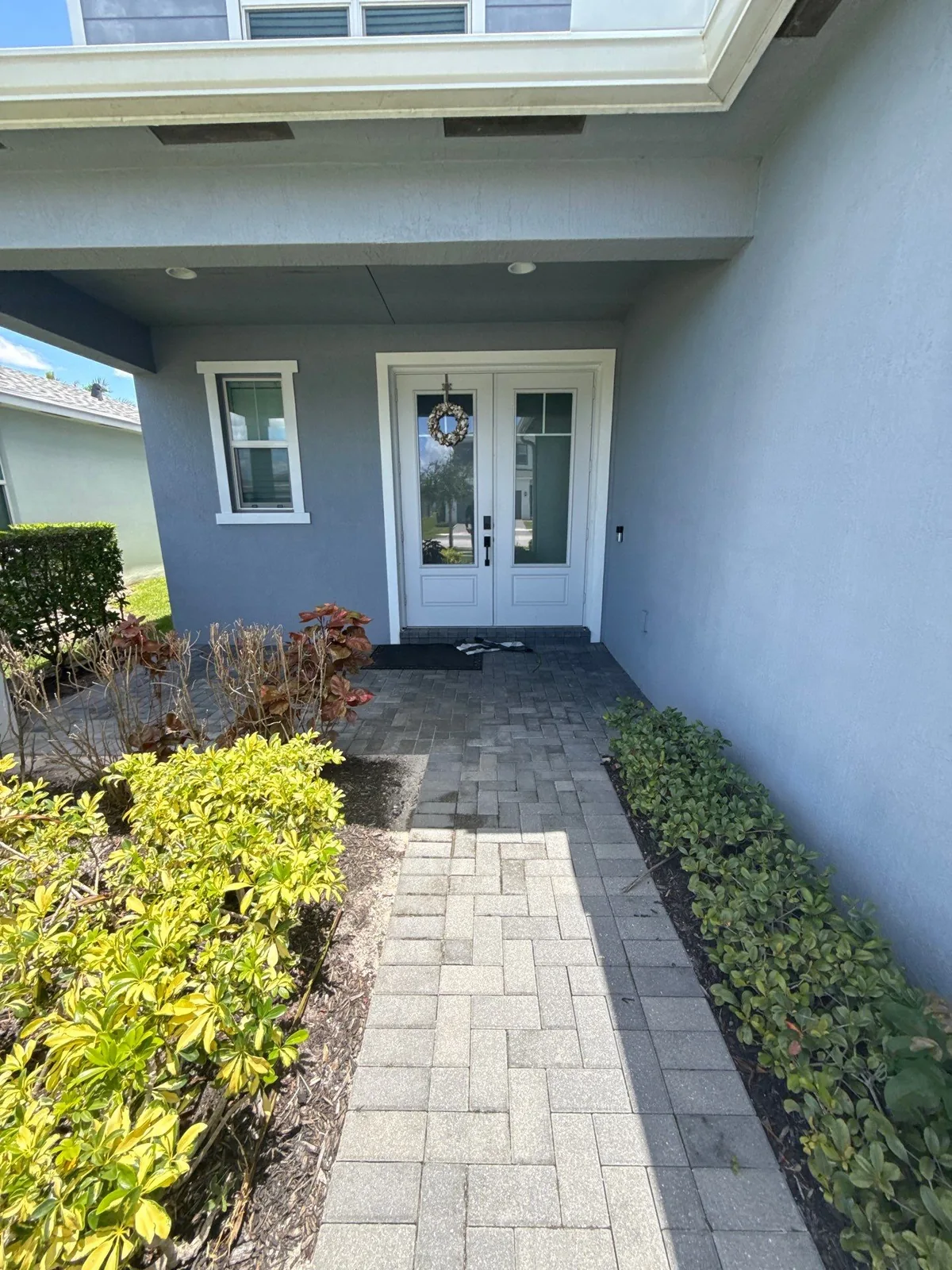 Exterior Painting in Port Saint Lucie, Florida. Front entry of a grey-blue stucco home with white double doors, white window trim and a paver walkway between hedges.