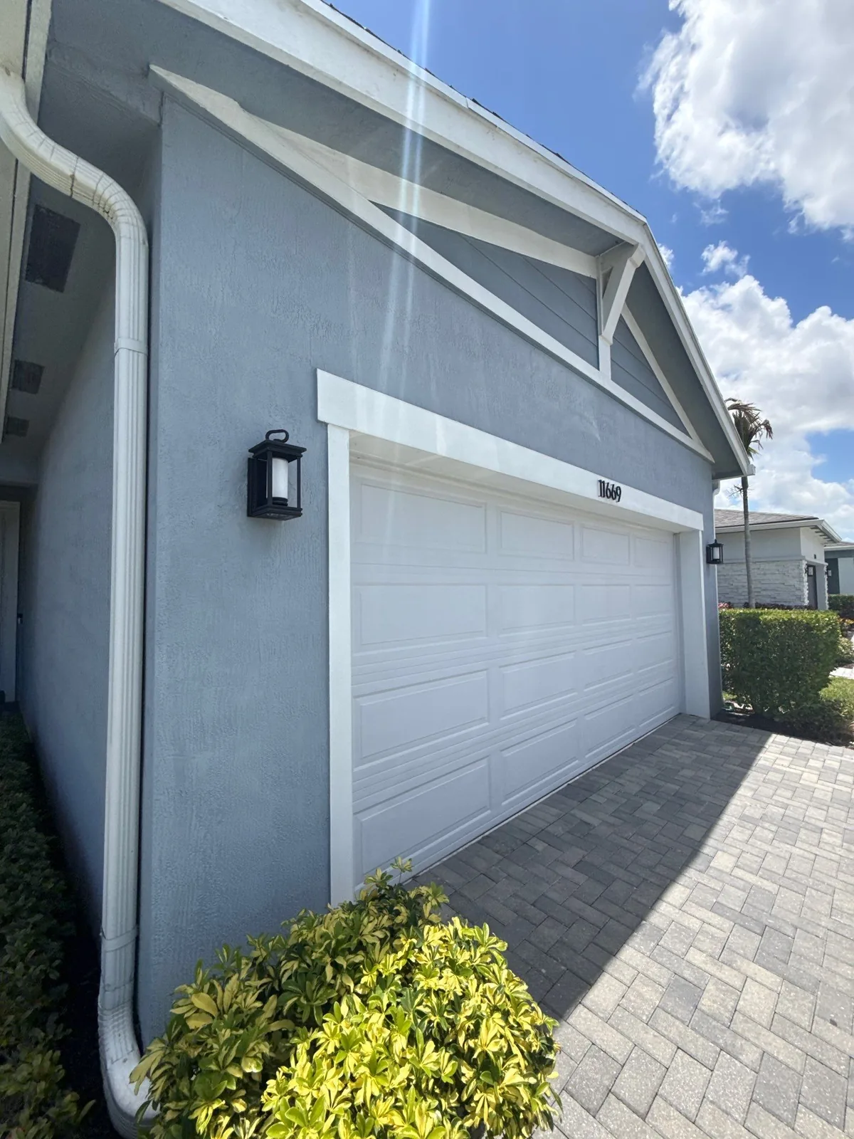 Exterior Painting in Port Saint Lucie, Florida. Attached two-car garage with grey stucco, white garage door, white gable trim, black coach light and paver driveway.