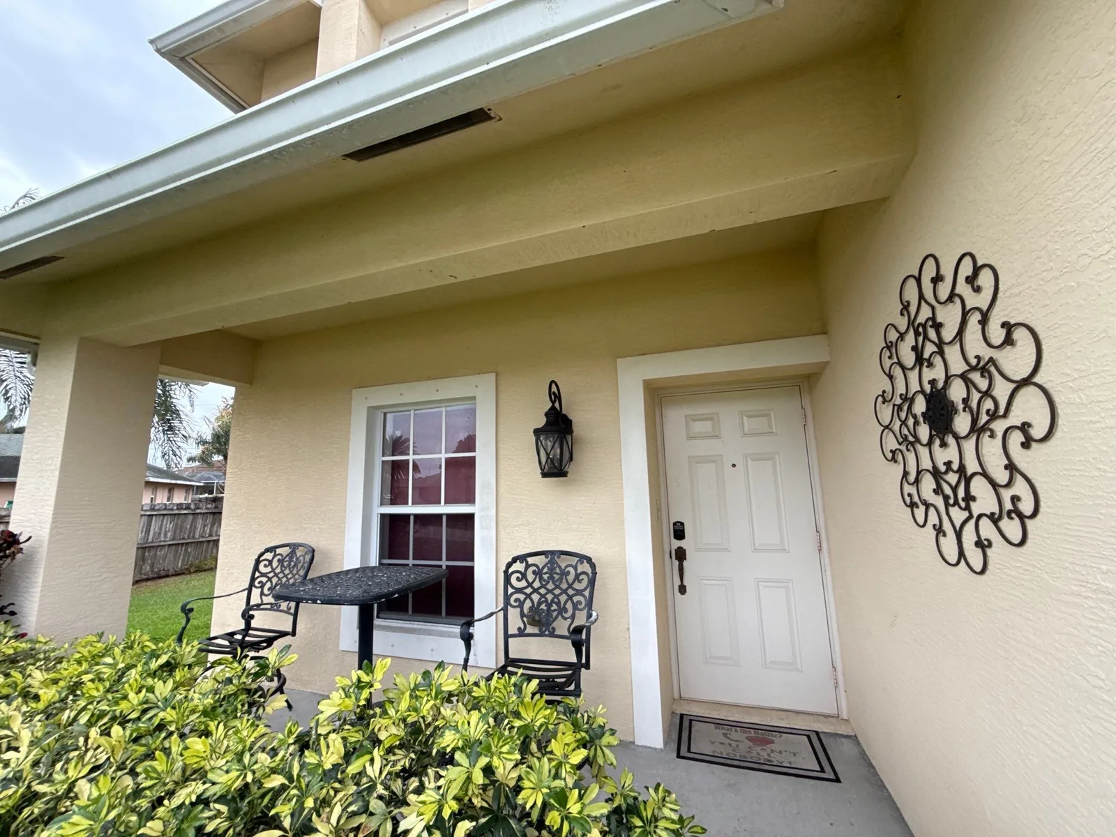 Exterior Painting in Port Saint Lucie, Florida. Covered front entry with cream stucco walls, white front door and trim, and black metal chairs.