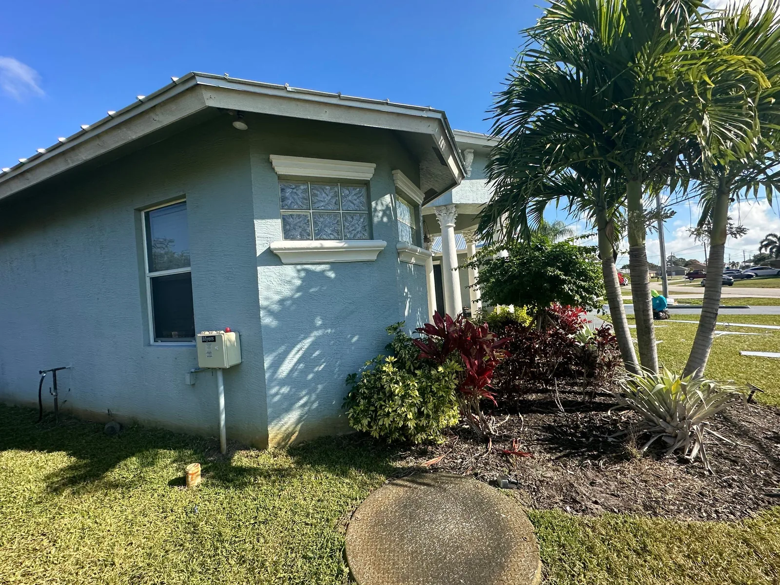 Exterior Painting in Port Saint Lucie, Florida. Single-storey home exterior with blue-grey stucco walls and white trim, palm trees and shrubs in front, under a clear blue sky.