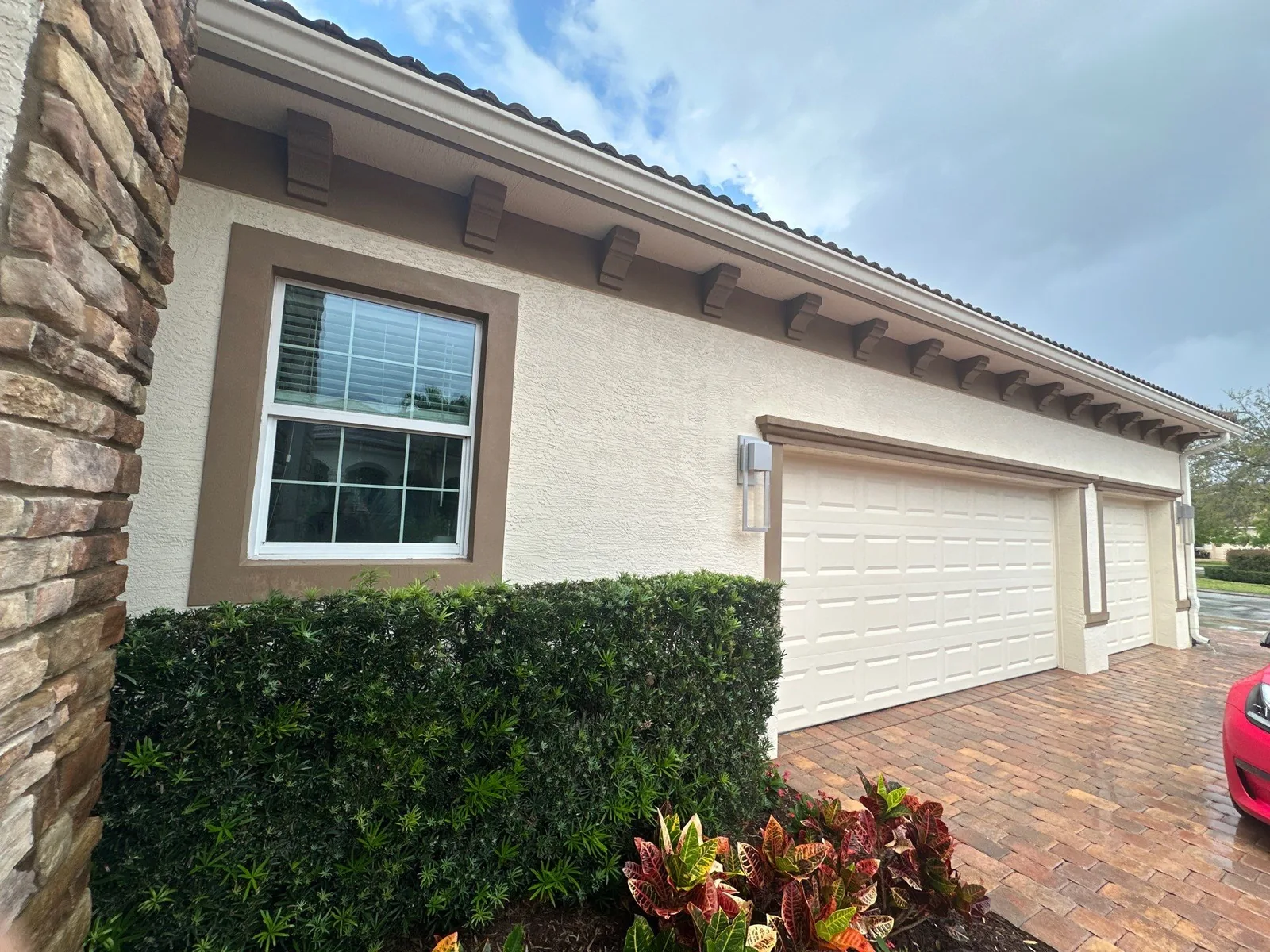 Exterior Painting in Port Saint Lucie, Florida. Single-storey stucco home exterior in beige with brown trim and fascia corbels, white garage doors, stone veneer corner and clipped hedge.