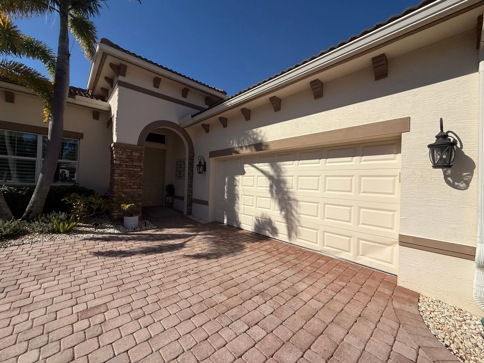 Exterior Painting in Port Saint Lucie, Florida. Front of a single-storey stucco home in cream with a tile roof, arched entry and two-car garage door, brick paver driveway.