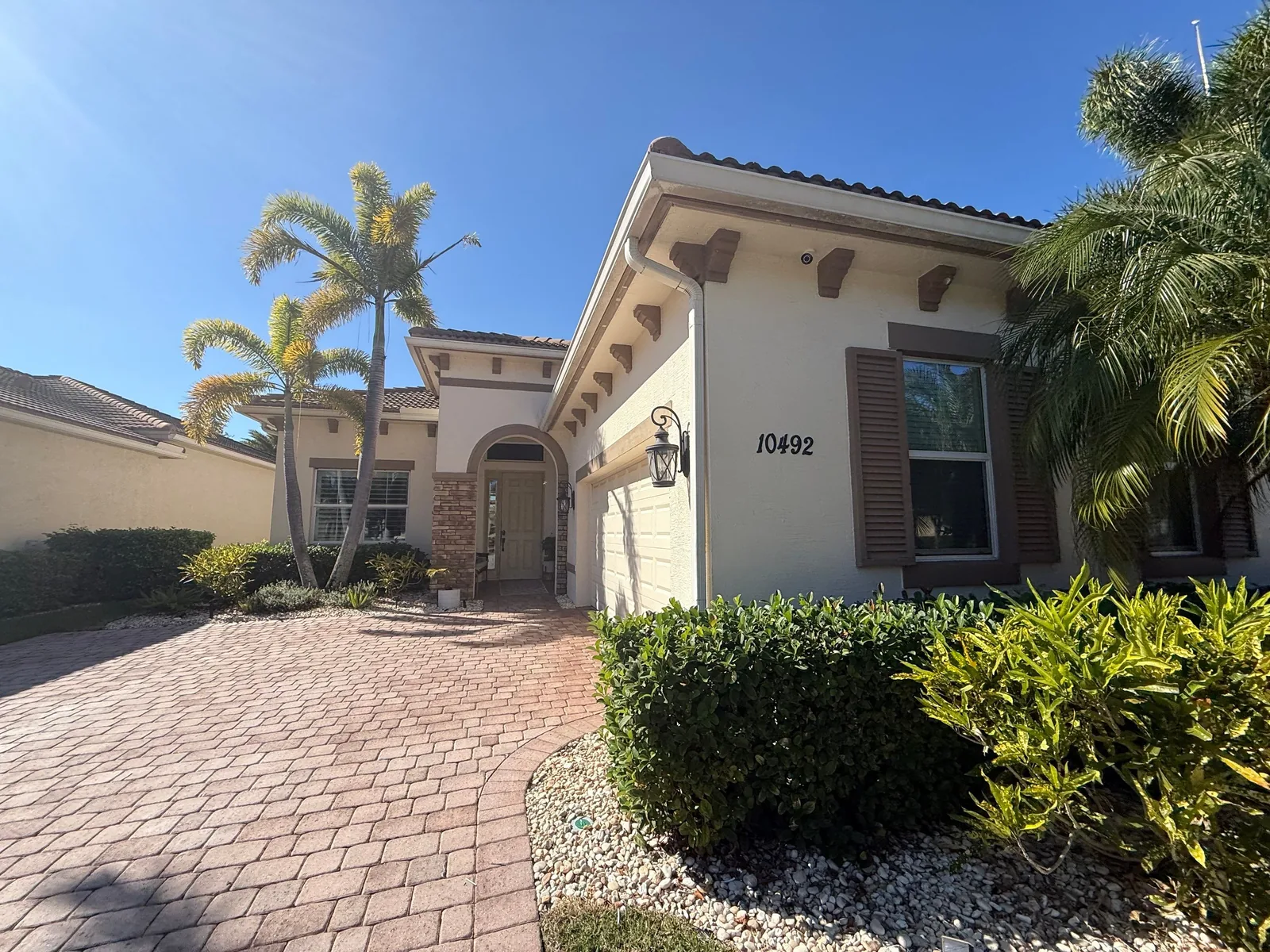 Exterior Painting in Port Saint Lucie, Florida. Front elevation of a cream stucco home with tile roof, palm trees and trimmed hedges along a paver driveway.